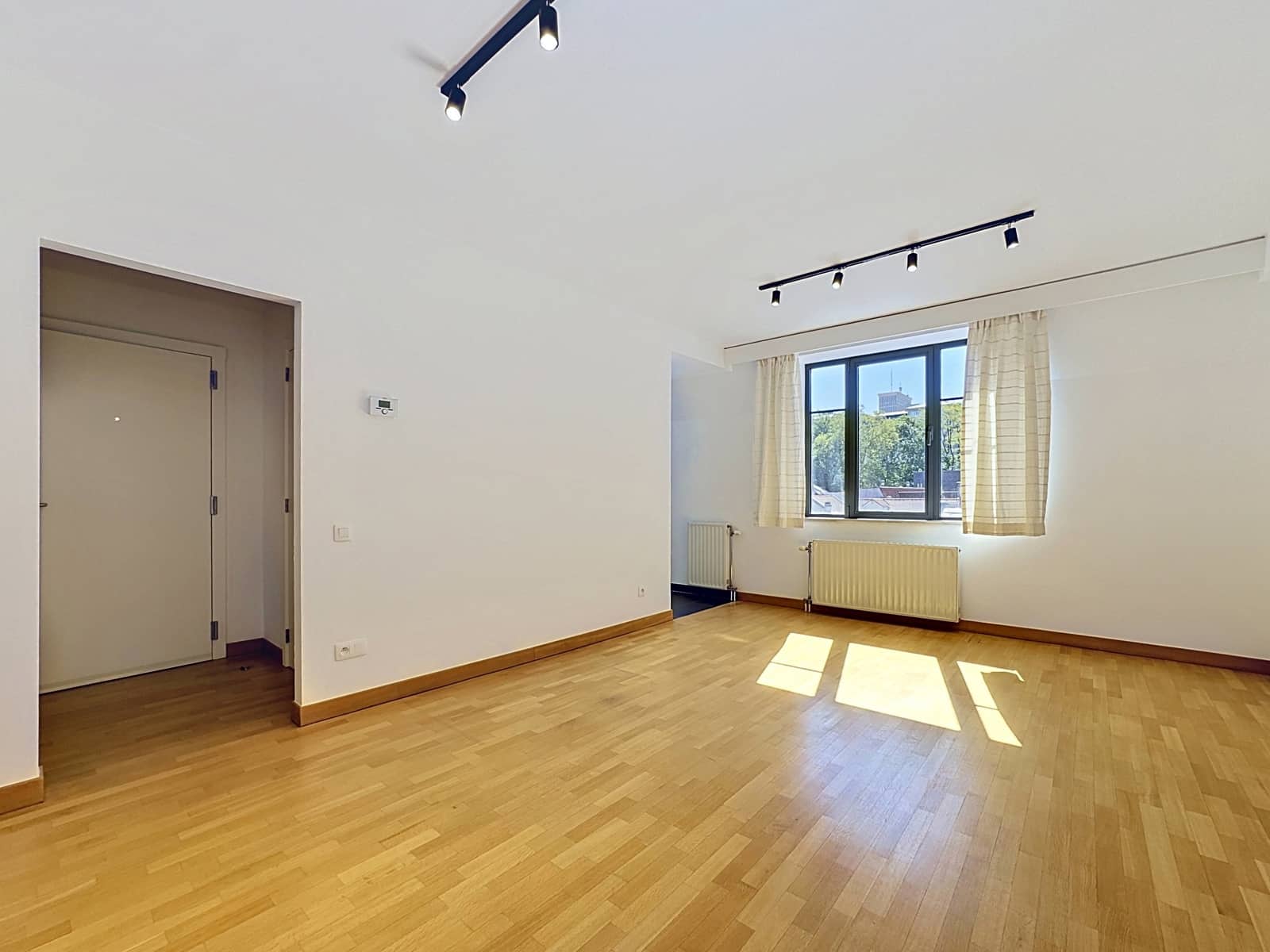 whise photo 2 Bright empty living room with wooden floor, white walls, a window with cream curtains, and ceiling track lighting.