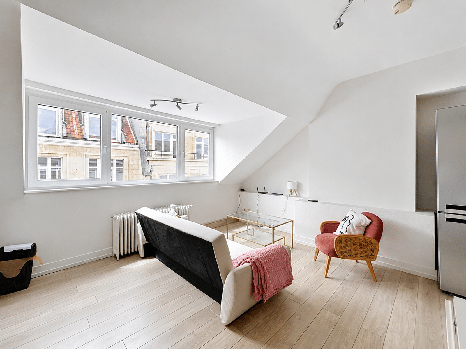 whise photo 2 Bright attic living room with a cream sofa, pink blanket, glass coffee table, and a red woven chair under a large angled window.