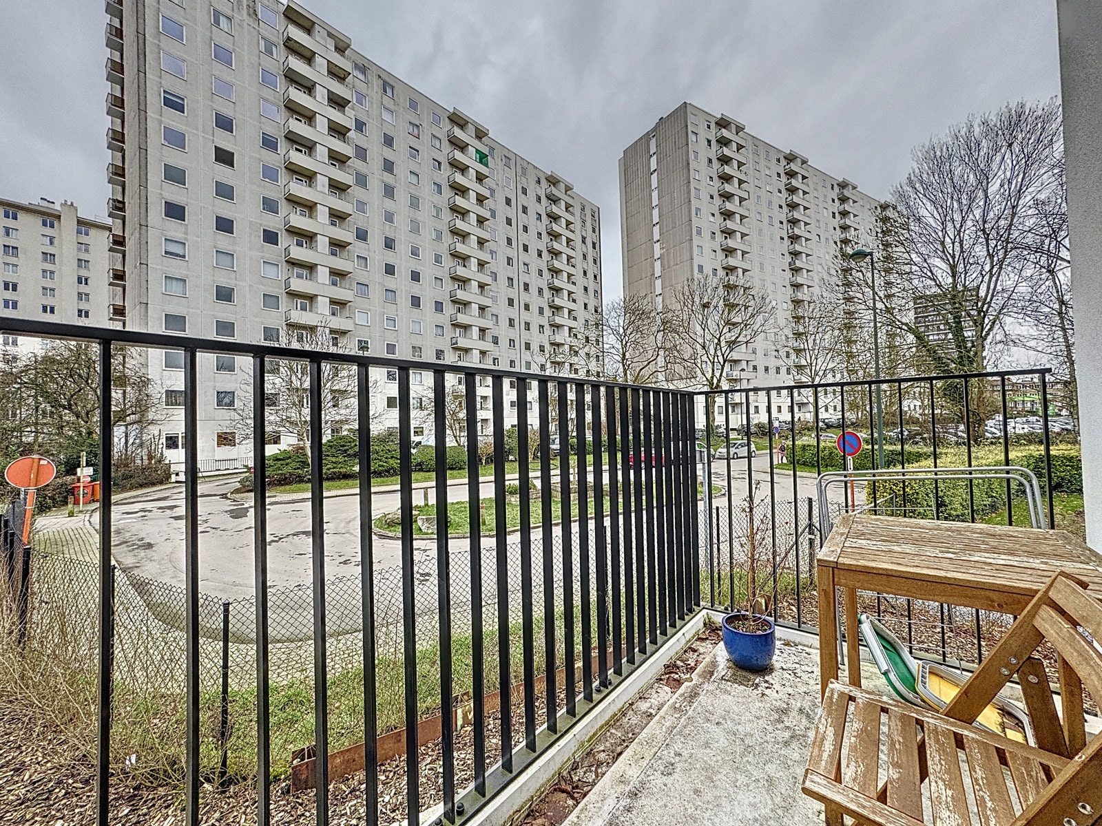 whise photo 10 Balcony with a wooden table and notebook-style chair, overlooking a gray apartment complex with many windows and balconies under a cloudy sky.