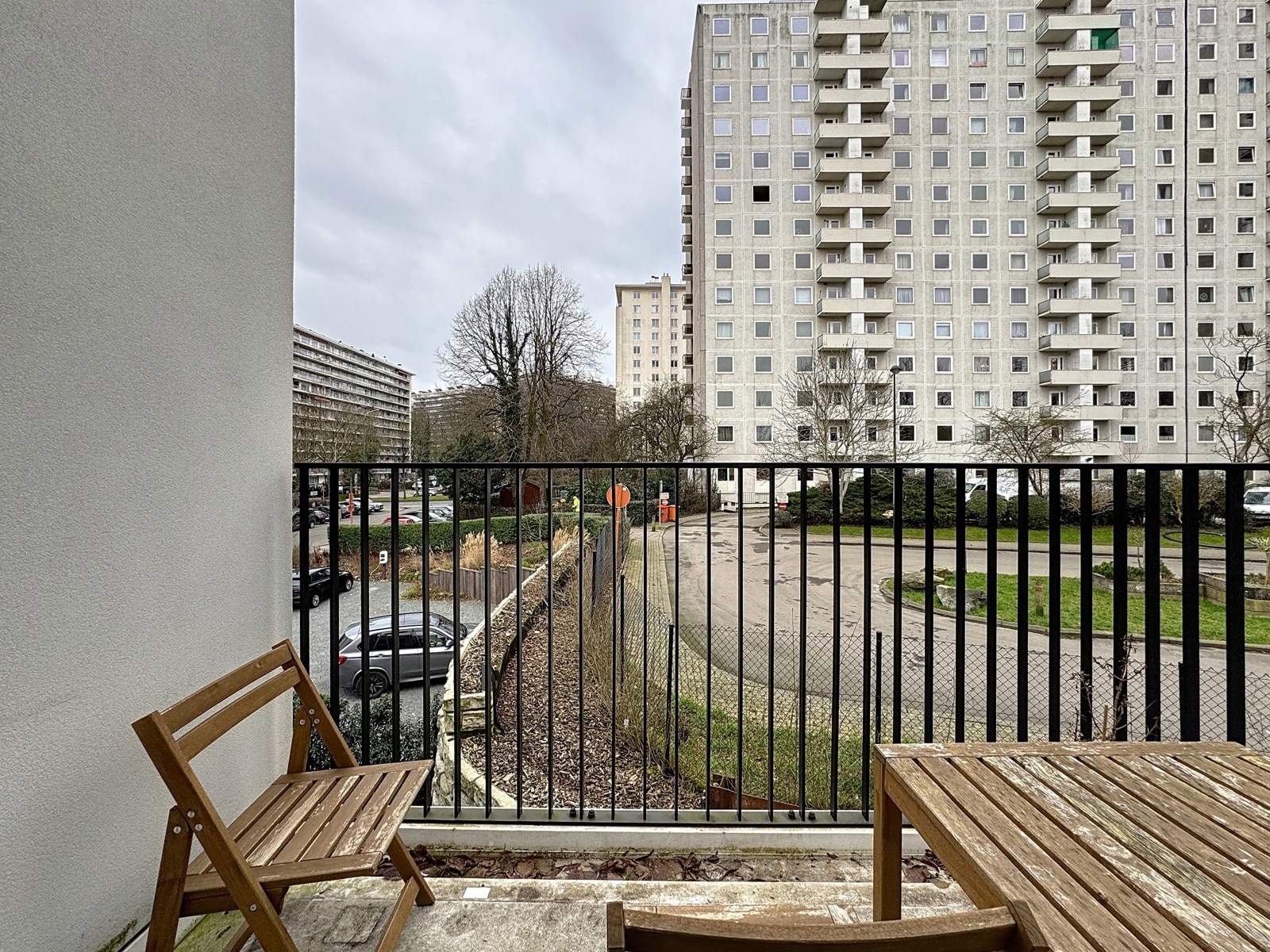 whise photo 9 View from a balcony with wooden chairs and table, overlooking a courtyard and tall grey apartment buildings on an overcast day.