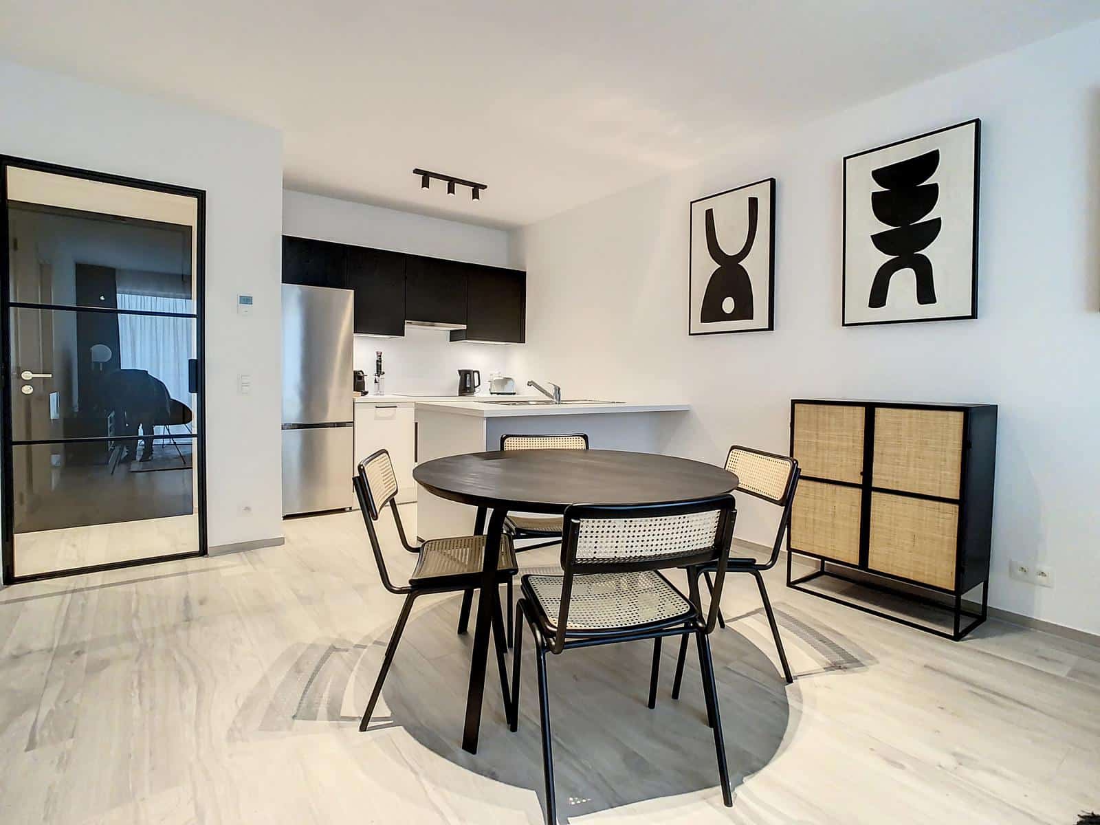 whise photo 2 Open-plan kitchen and dining area with a round dark table and four cane chairs, white walls, and black upper cabinets.