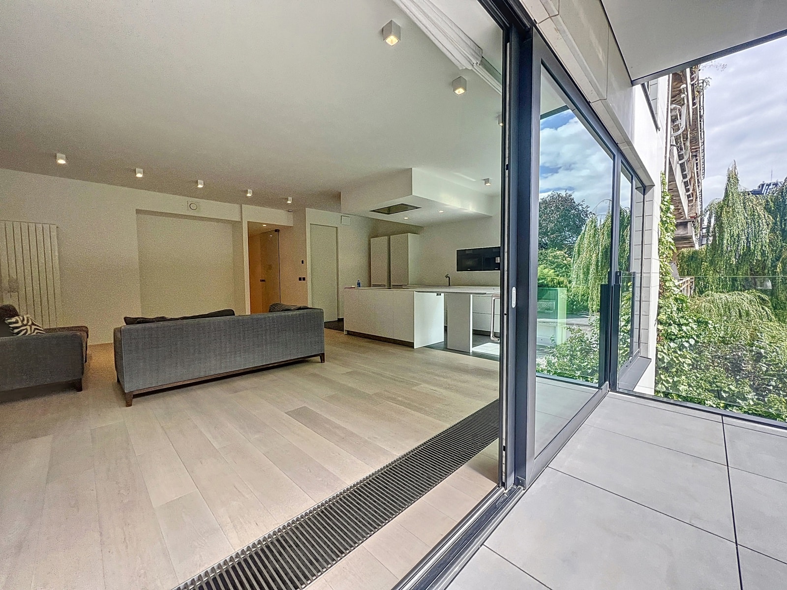 whise photo 3 Open-concept living area with gray sofas facing a white kitchen island, and large glass doors opening to a sunlit patio outside.