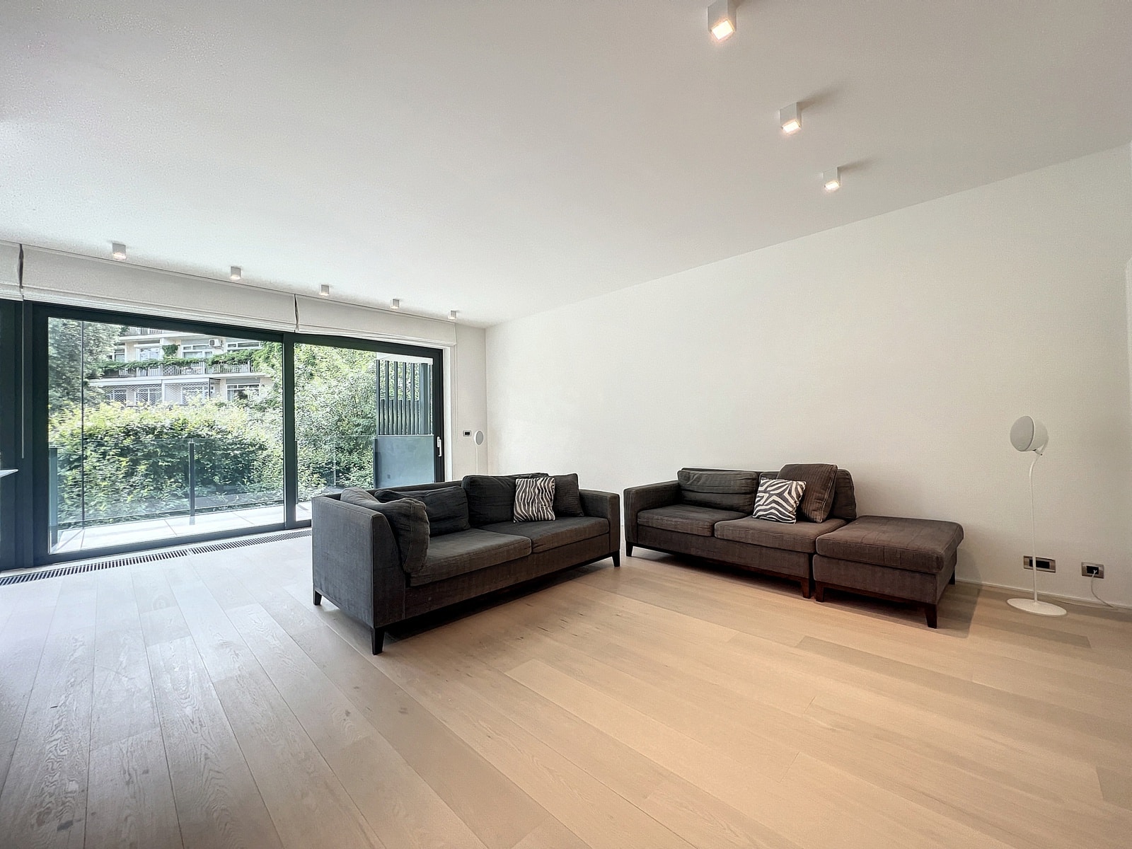 whise photo 10 Bright, minimalist living room with light wood floors, two dark gray sofas with zebra-print cushions, and large glass sliding doors opening to a balcony.