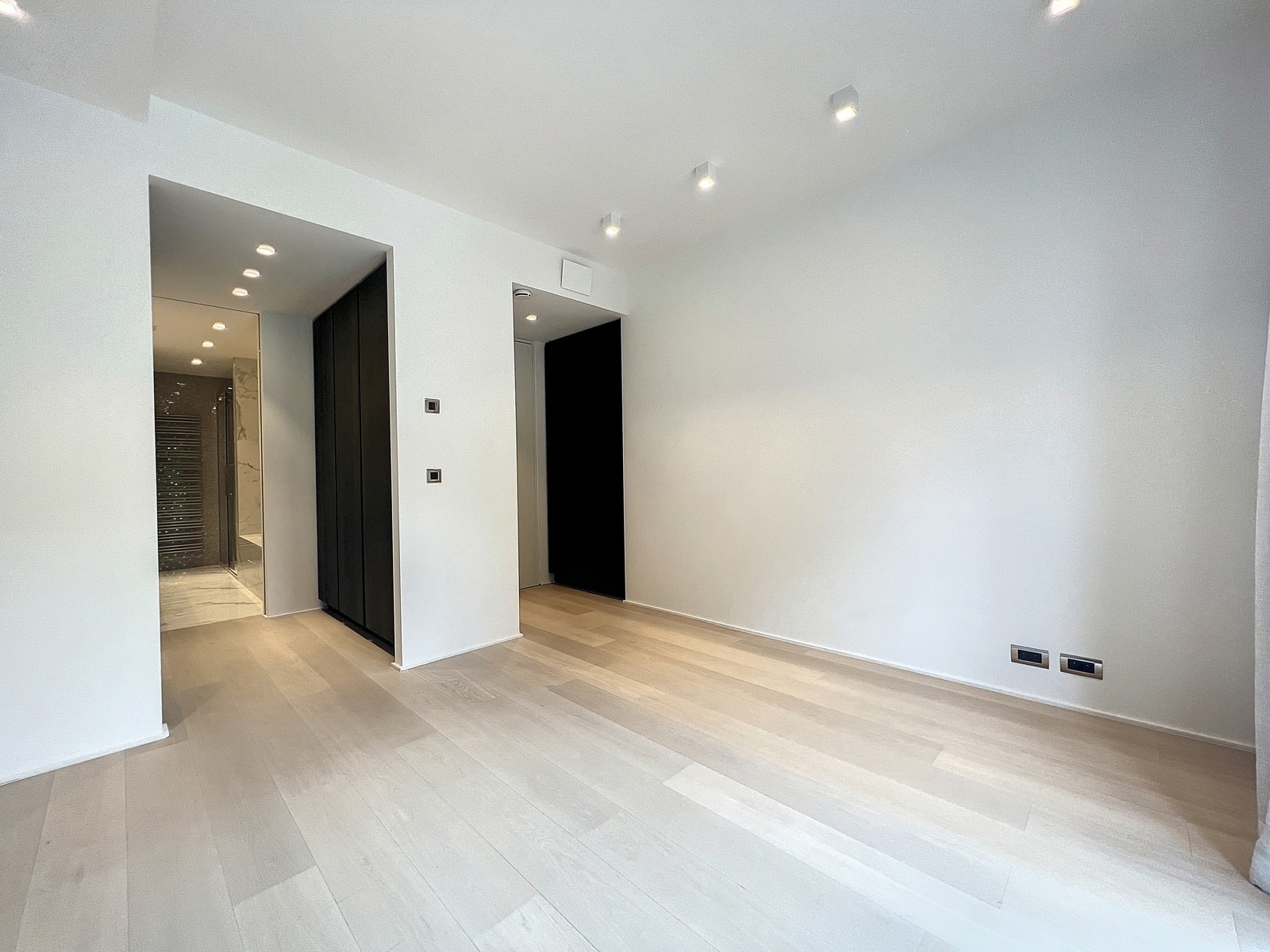 whise photo 12 Bright, empty modern living area with white walls, light wood floors, and ceiling spotlights; doorway leads to a marble-tiled bathroom.