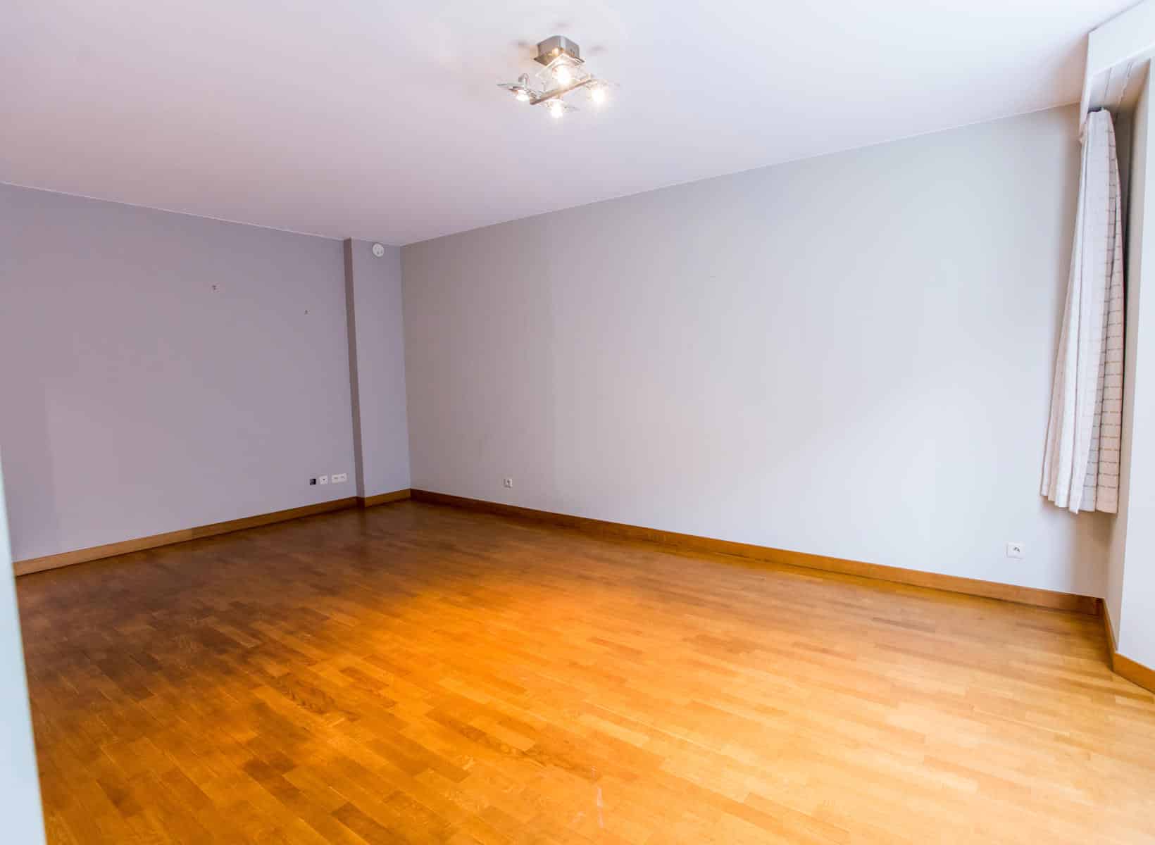 whise photo 3 Empty rectangular room with light gray walls, wooden floor, and a modern ceiling light fixture; window with beige curtain on the right.