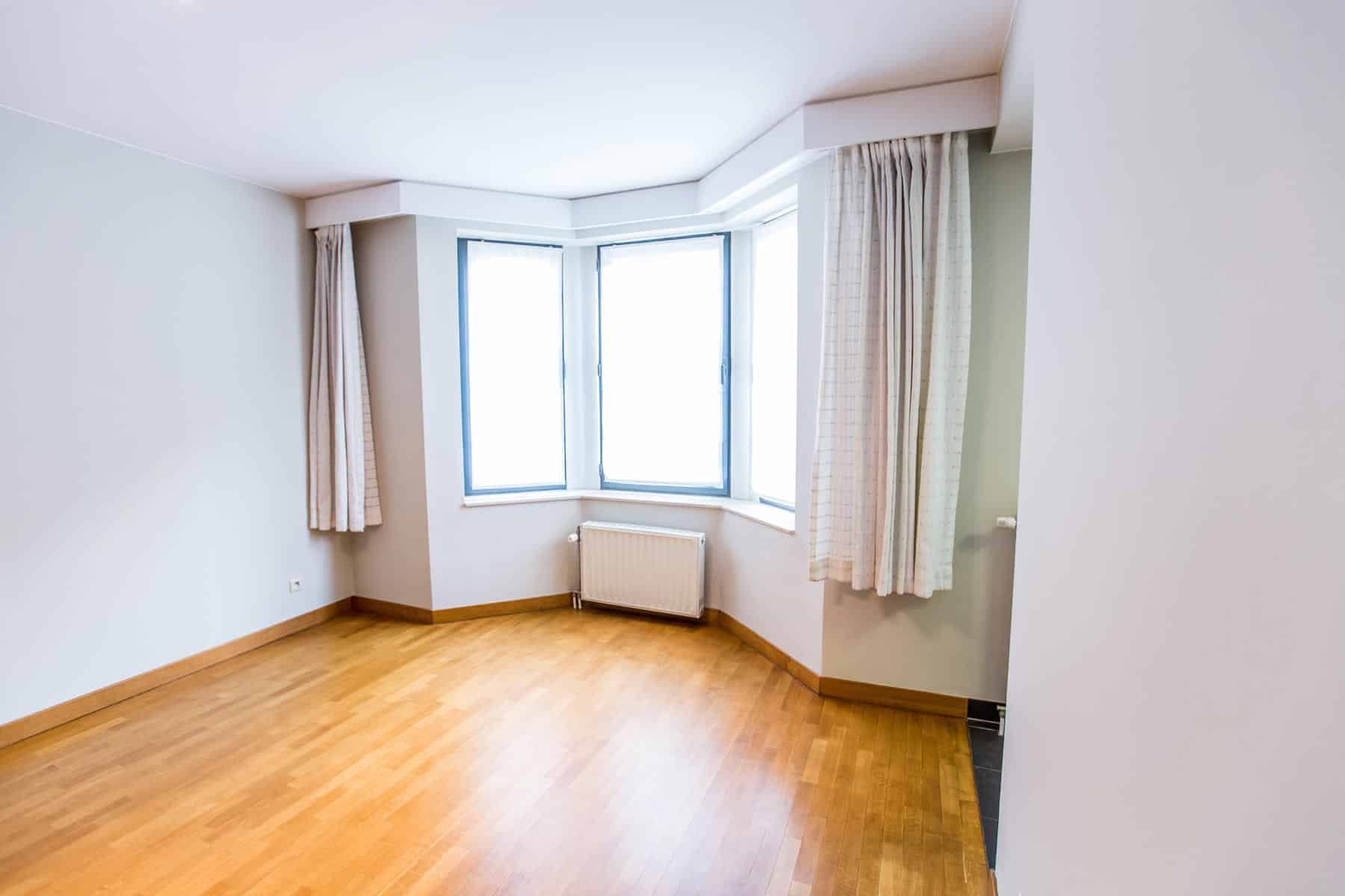 whise photo 2 Empty room with light wood flooring and a bay window framed by white trim and beige curtains, radiator beneath the window.