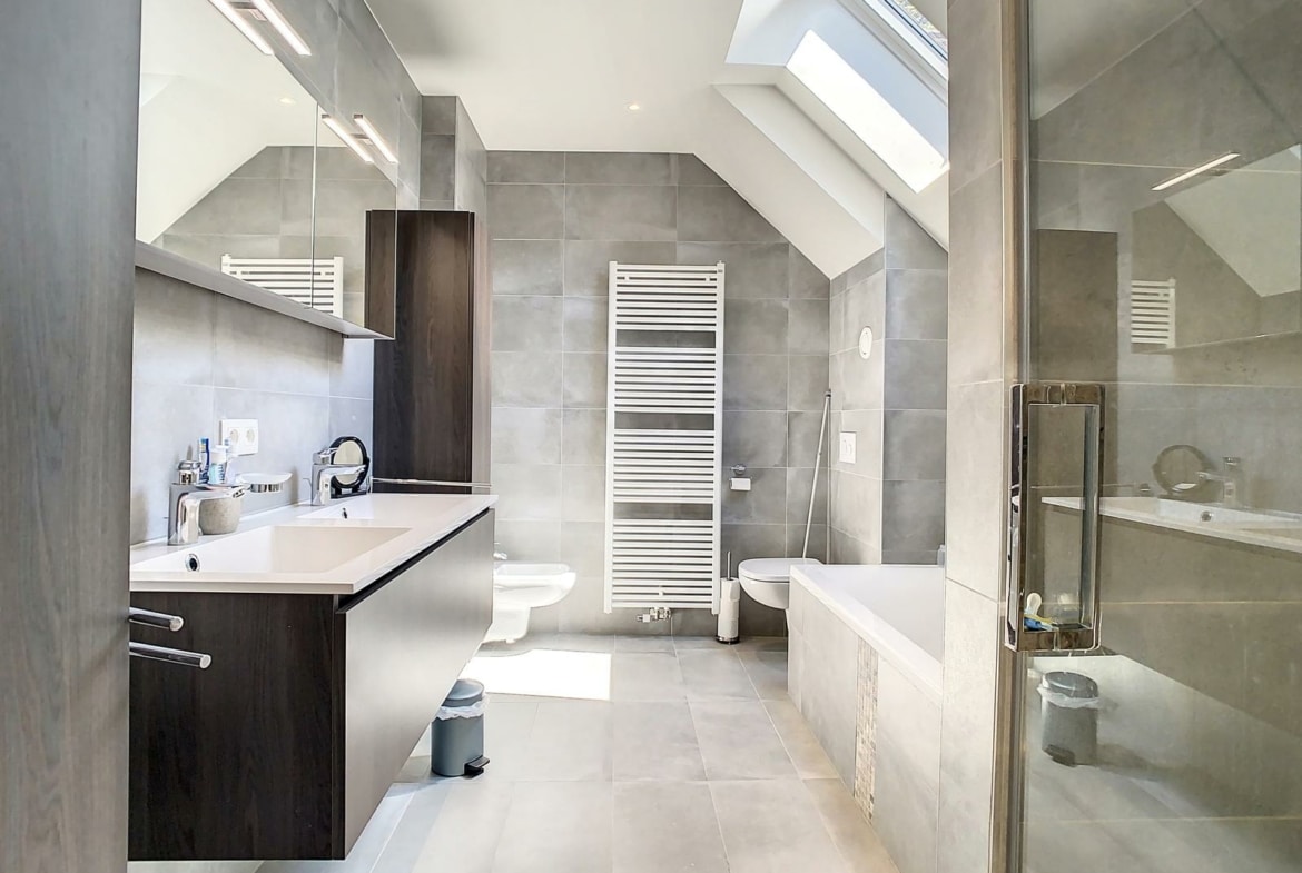 Modern gray-tiled bathroom with a skylight, wall mirror, double sink vanity, white heated towel rail, toilet, bidet, glass shower and bathtub.