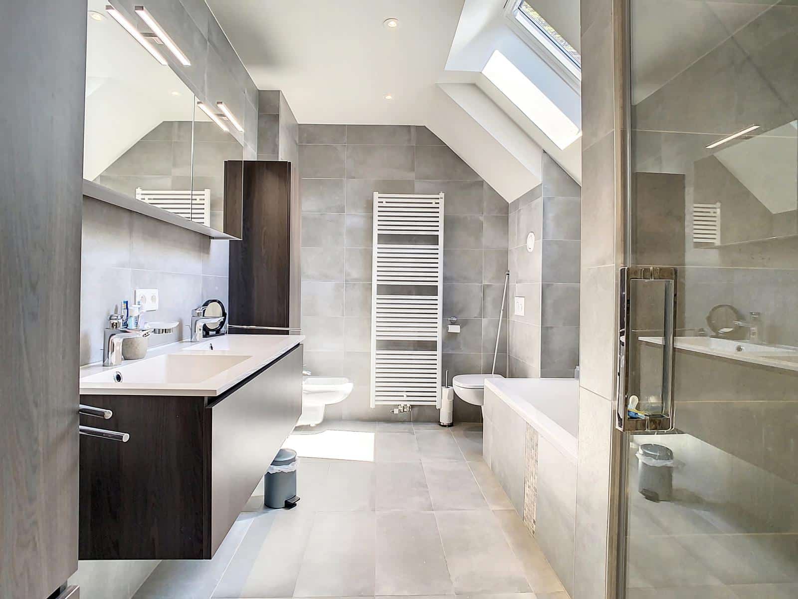 Wijze foto 13 Moderne badkamer met grijze tegels, voorzien van een dakraam, een wandspiegel, een dubbele wastafel, een wit verwarmd handdoekenrek, een toilet, een bidet, een glazen douchecabine en een bad.