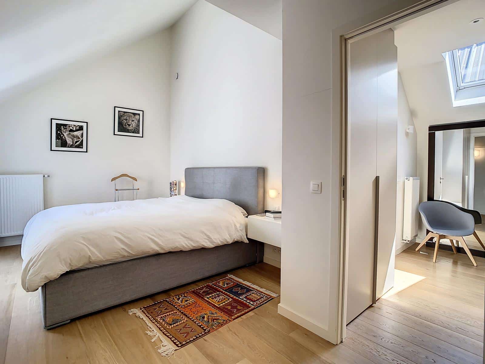 wijs foto 12 Minimalistische slaapkamer met witte muren en een lichte houten vloer, een grijs gestoffeerd bed, wit beddengoed, twee kleine nachtkastjes en een tapijt met patroon aan het voeteneinde van het bed.