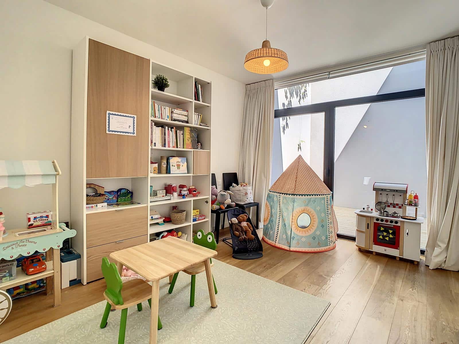 Wijze foto 10 "Een lichte speelkamer voor kinderen met een grote kast in wit en hout, een speelgoedkeuken en een speeltent van stof met een patroon, vlak bij een glazen deur."