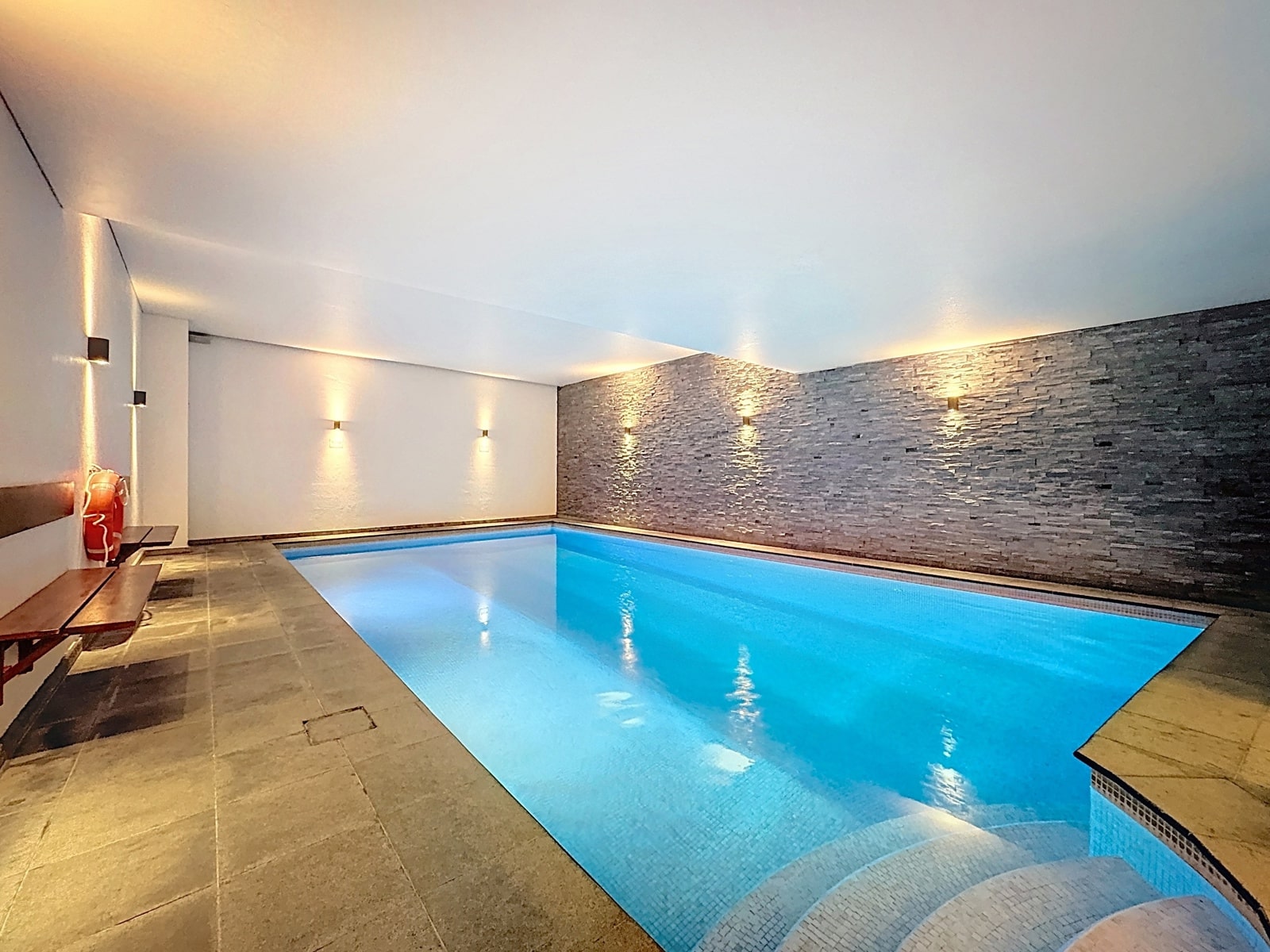 Wijze foto 8 Indoor swimming pool with blue water, gray tile deck, and a textured stone accent wall with warm wall lights.
