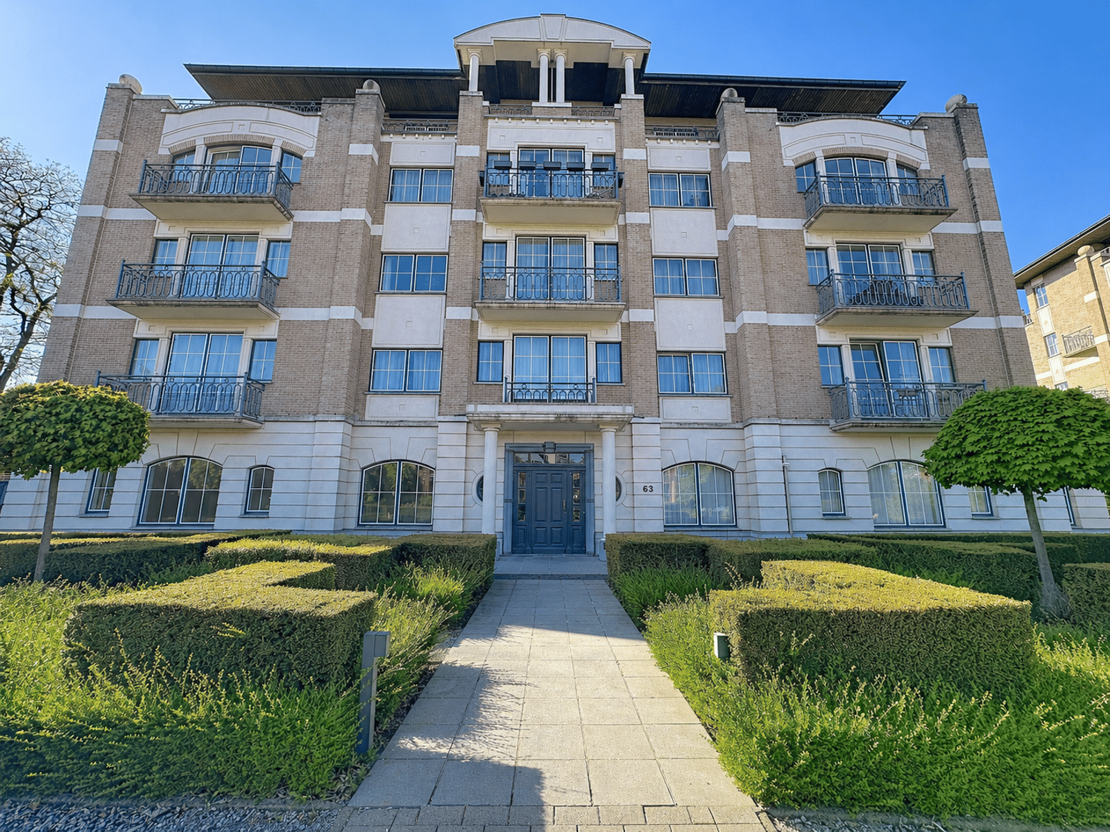 wijs foto 12 Front view of a brick apartment building with balconies, central blue door entrance, and a manicured hedge-lined walkway leading to it.