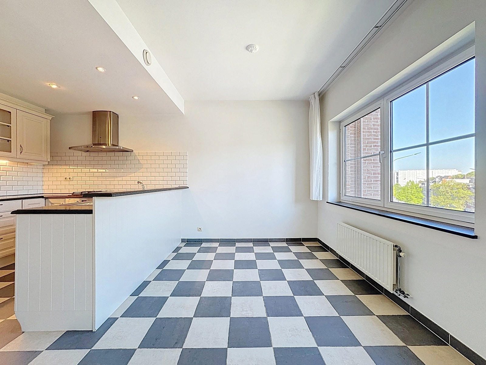 foto 5 Bright modern kitchen with black-and-white checkered floor, white cabinets, and a curved island with dark countertop beneath a stainless range hood; large window view to buildings outside.