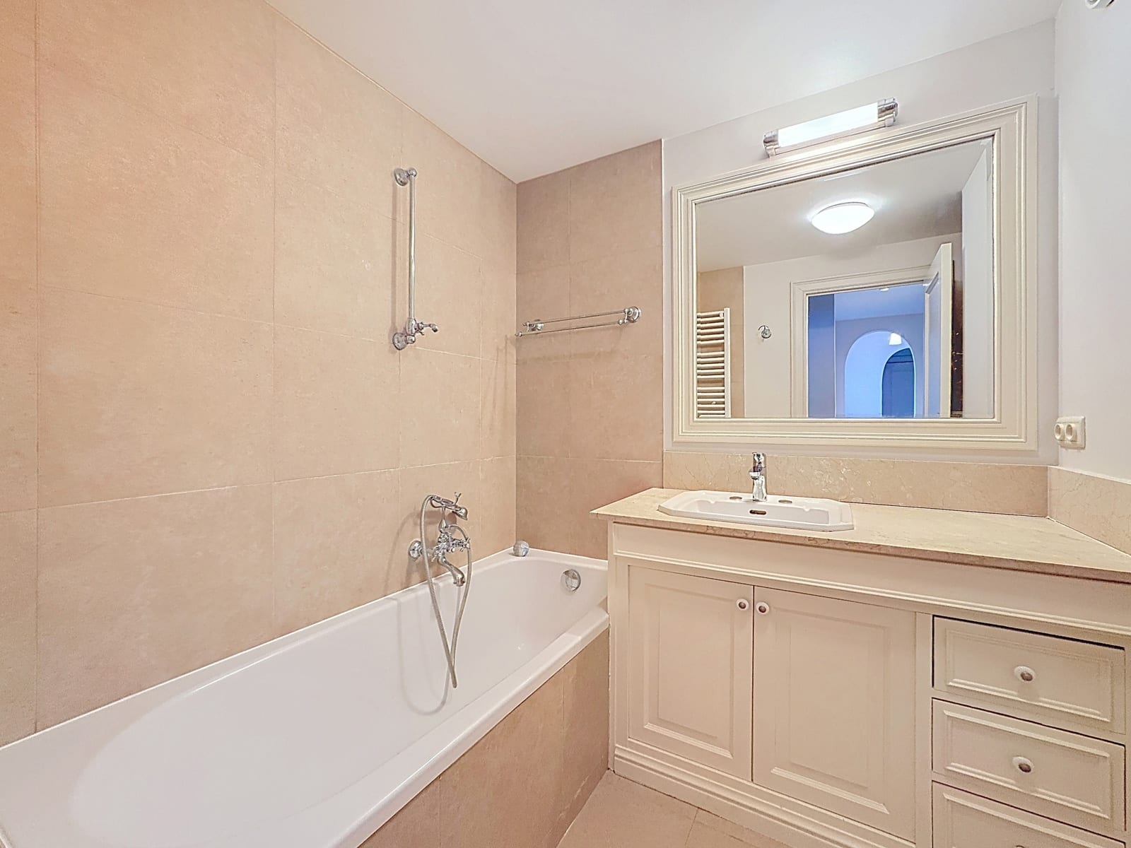 foto 6 Beige-tiled bathroom featuring a white bathtub, chrome faucet, towel bar, and a white vanity with a marble countertop under a large framed mirror.