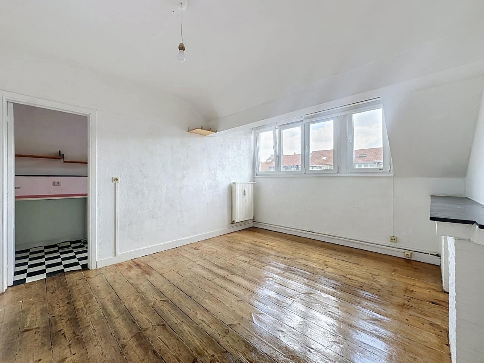 foto 2 Een lichte, lege zolderachtige kamer met witte muren, een rij deuren en ramen langs één muur, een radiator en een houten vloer; door een open deur is een kleine nis te zien met een zwart-wit geruite vloer.