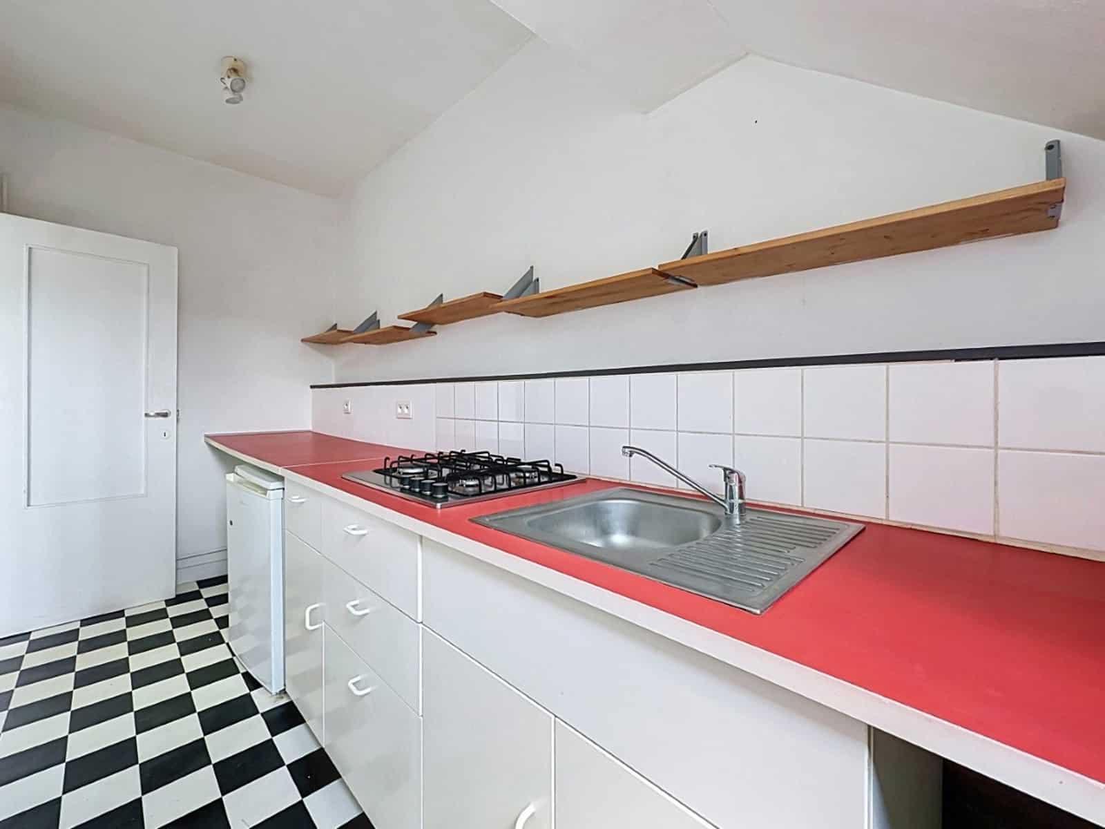 foto 4 Keuken met een rood aanrechtblad, een gasfornuis en een metalen spoelbak onder witte keukenkastjes; langs de muur boven het aanrecht loopt een houten plank.