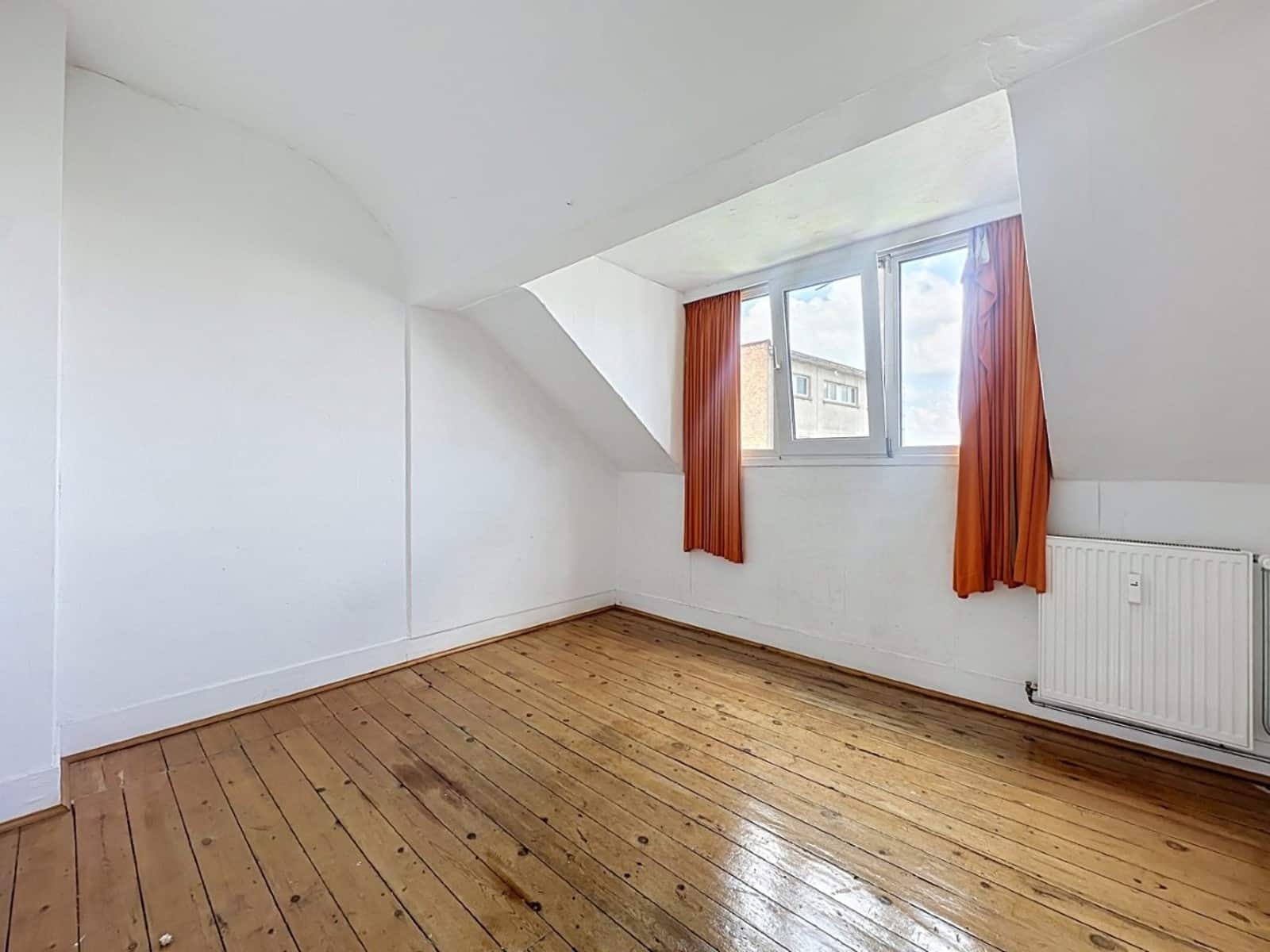 foto 5 Kleine, lege zolderkamer met witte schuine plafonds, een honingkleurige houten vloer en een raam met oranje gordijnen waardoor het daglicht naar binnen valt.