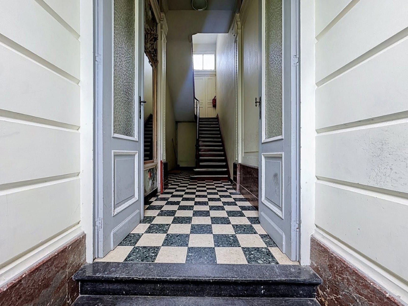 Wijze foto 8 Entreehal met een zwart-witte dambordvloer; de deuren geven toegang tot een trappenhuis boven. Het licht bovenaan zorgt voor perspectief richting de trap.