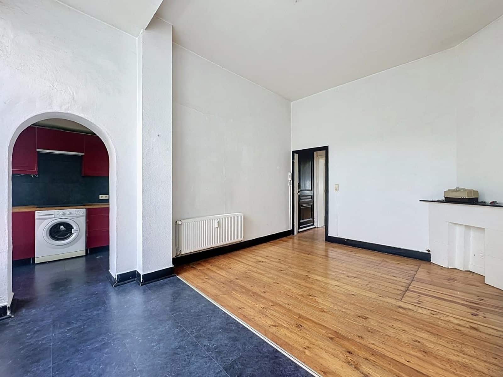 whise photo 2 Bright living room with mixed wood and dark tile flooring; arched doorway reveals a small kitchen with red cabinets and a washing machine nearby.
