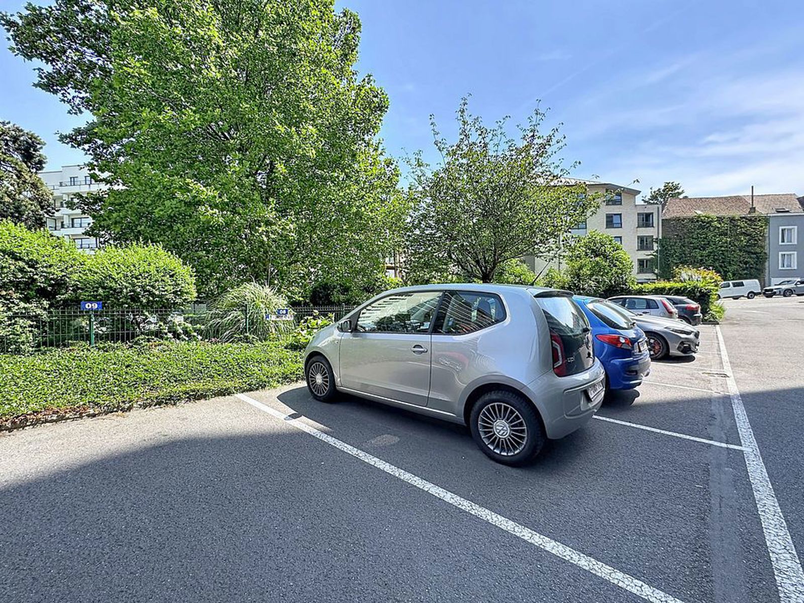 Parkeerterrein in de open lucht met bomen – You Real Estate Agency Een met bomen omzoomde parkeerplaats buiten bij een wooncomplex met appartementen en geparkeerde auto’s.