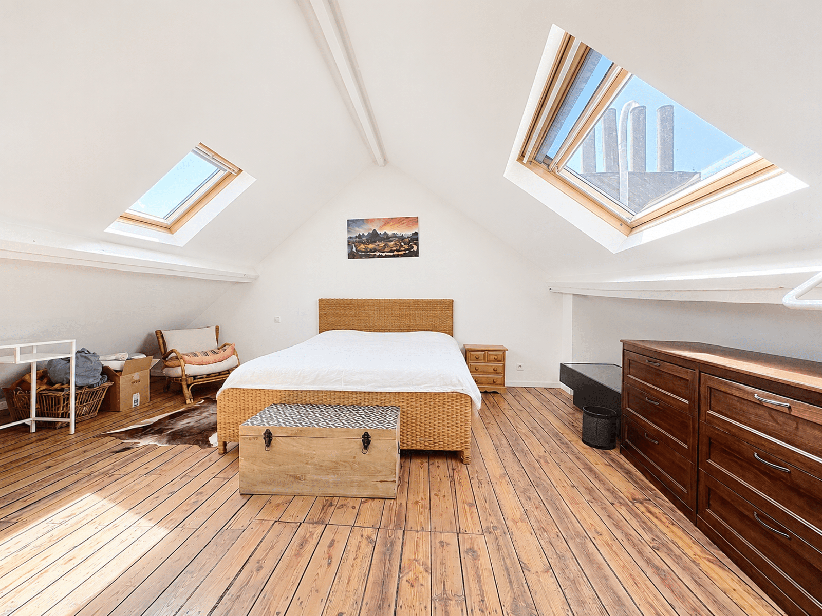 Chambre Mansardée Lumineuse – You Real Estate Agency Chambre mansardée lumineuse de maison avec parquet, lit en rotin et fenêtres de toit.