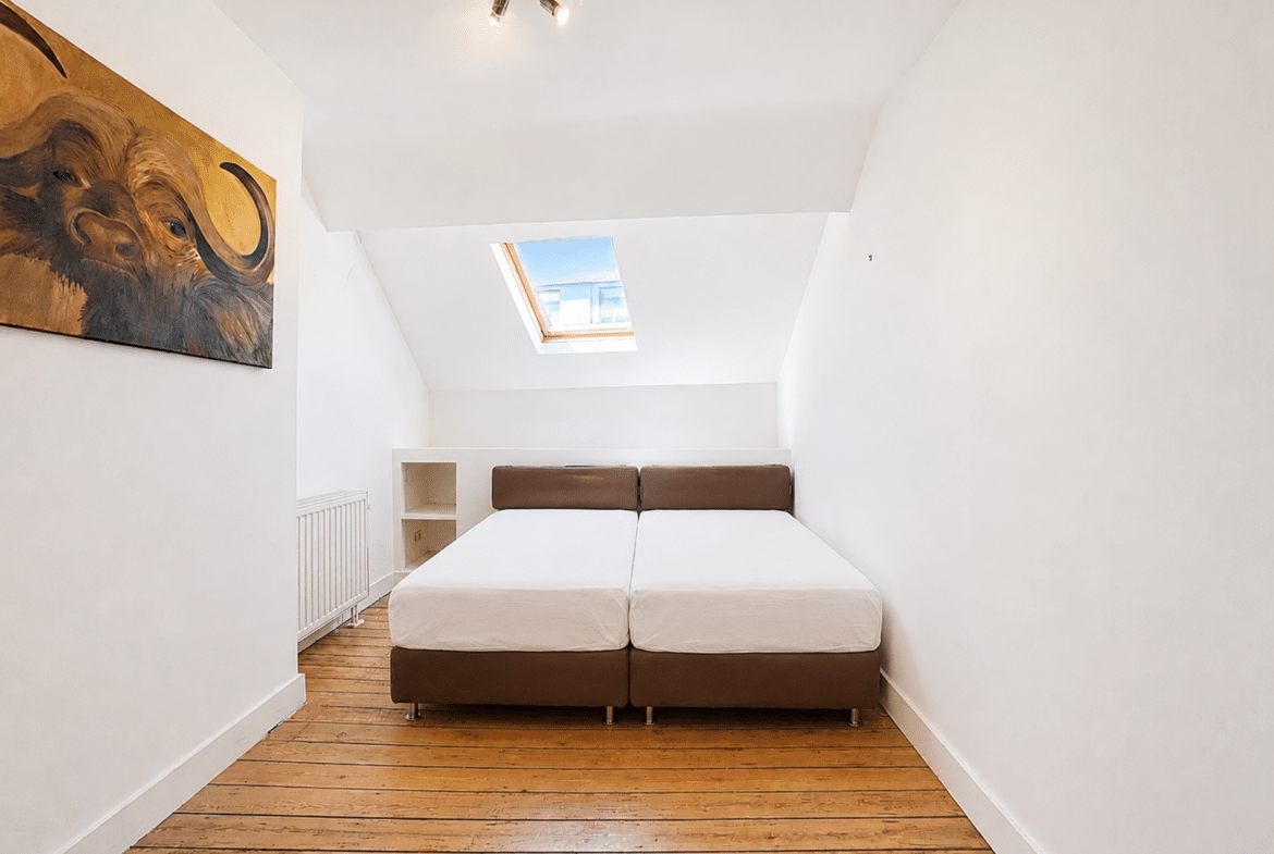 Chambre mansardée lumineuse avec deux lits, parquet et fenêtre de toit dans appartement.