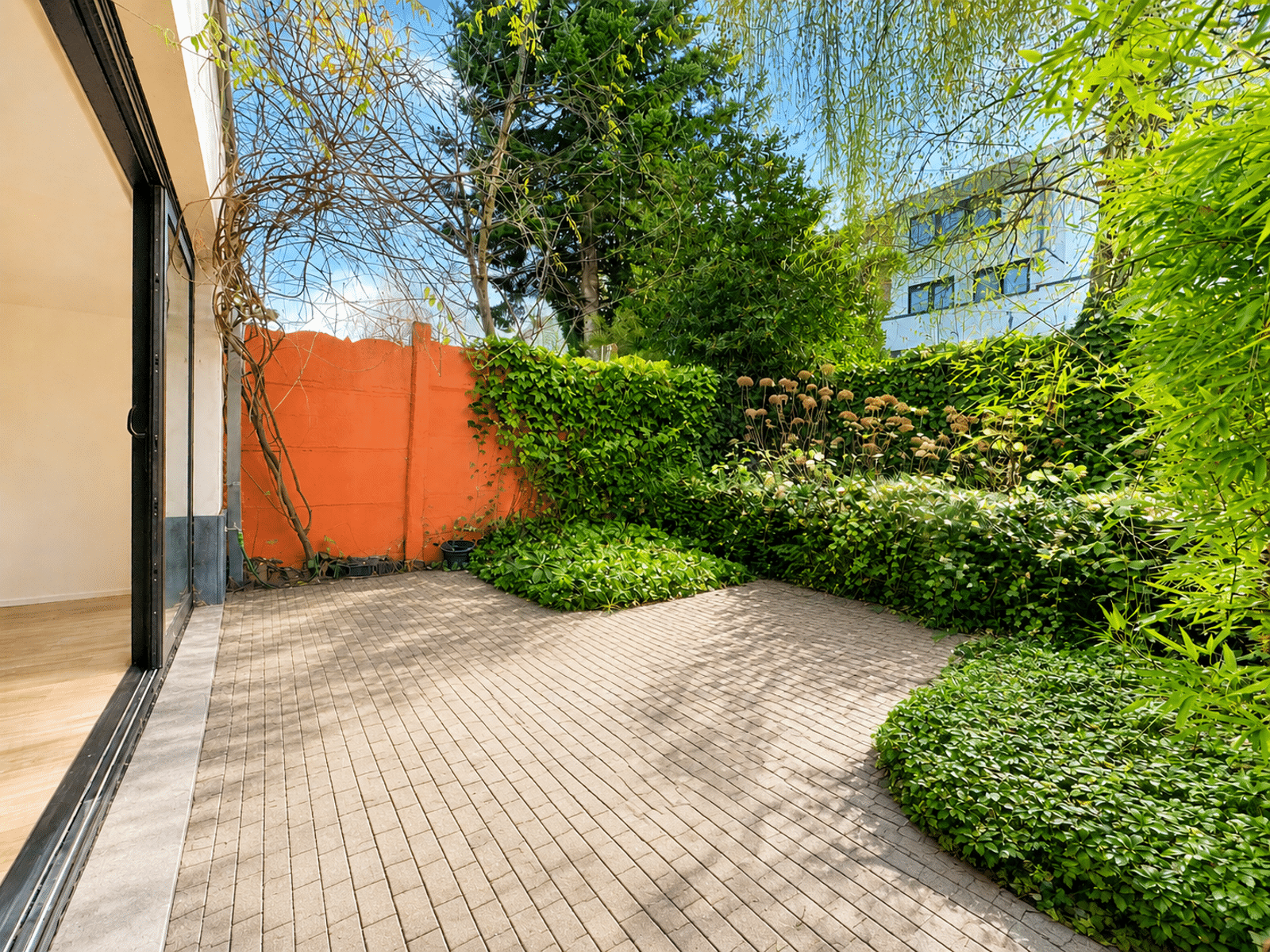 Patio Verdoyant avec Mur Orange – You Real Estate Agency Terrasse intérieure verdoyante d’une maison moderne avec baie vitrée et mur orange.