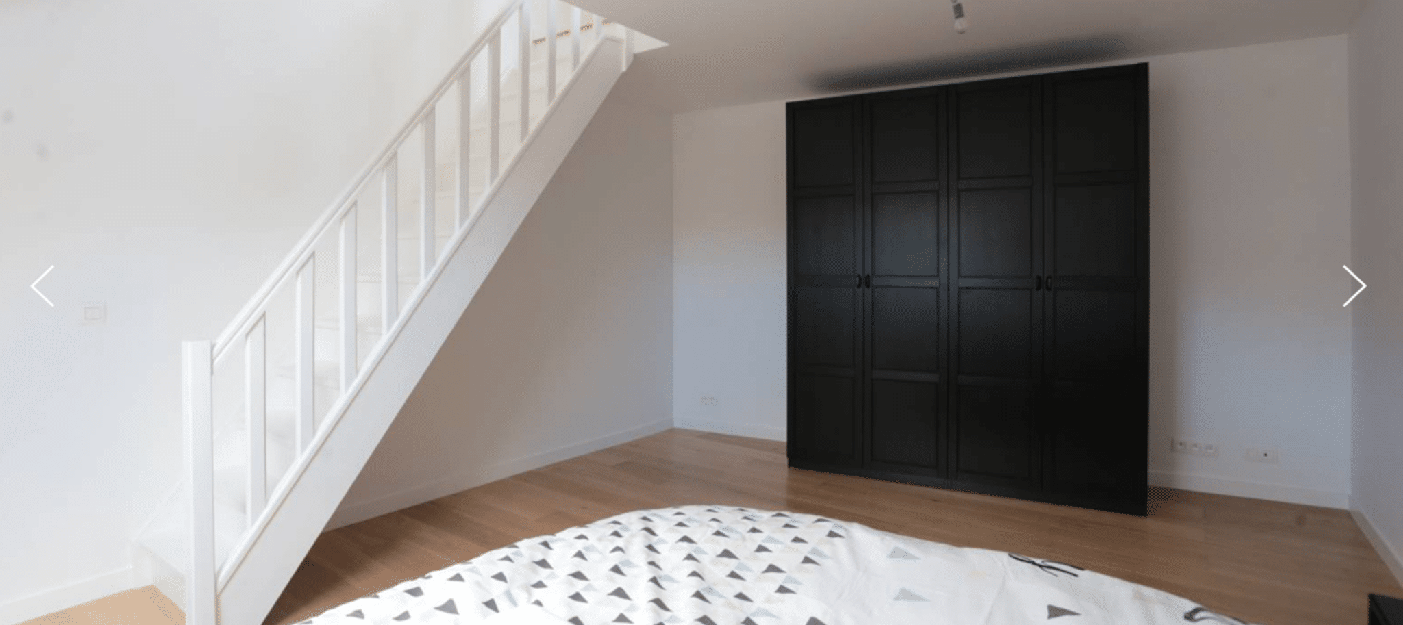 Chambre Lumineuse avec Escalier Blanc – You Real Estate Agency Chambre d’appartement lumineuse avec escalier blanc, parquet et grande armoire noire.
