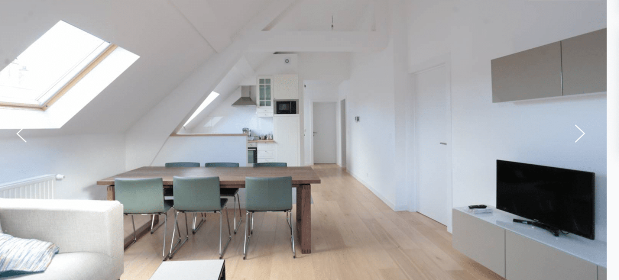 whise photo 2 Bright open-plan attic living area featuring a wooden dining table with teal chairs, a compact kitchen, and a wall-mounted TV.