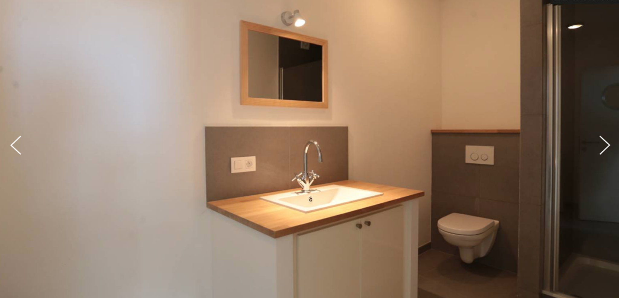 Salle de Bain Moderne – You Real Estate Agency Modern bathroom vanity with wood countertop, white sink, chrome faucet, and framed mirror; toilet visible on the right.