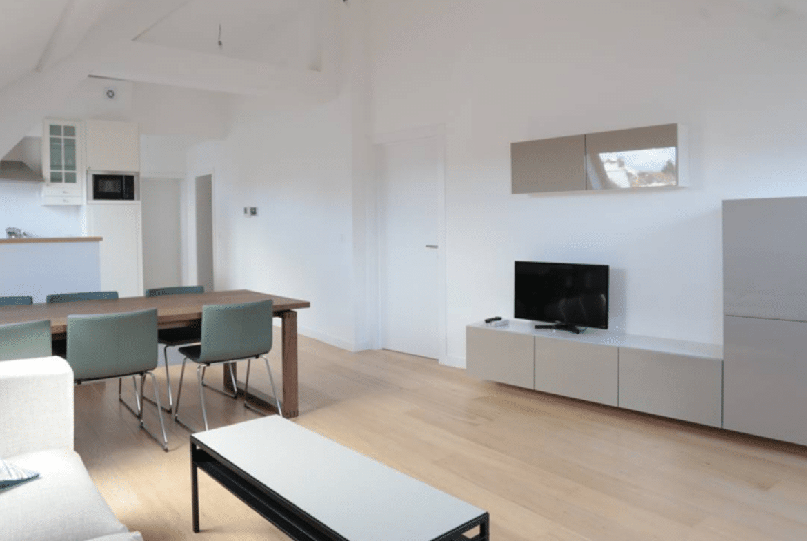 Open-plan living space with a wooden dining table and teal chairs, white walls, and a TV cabinet on the right.