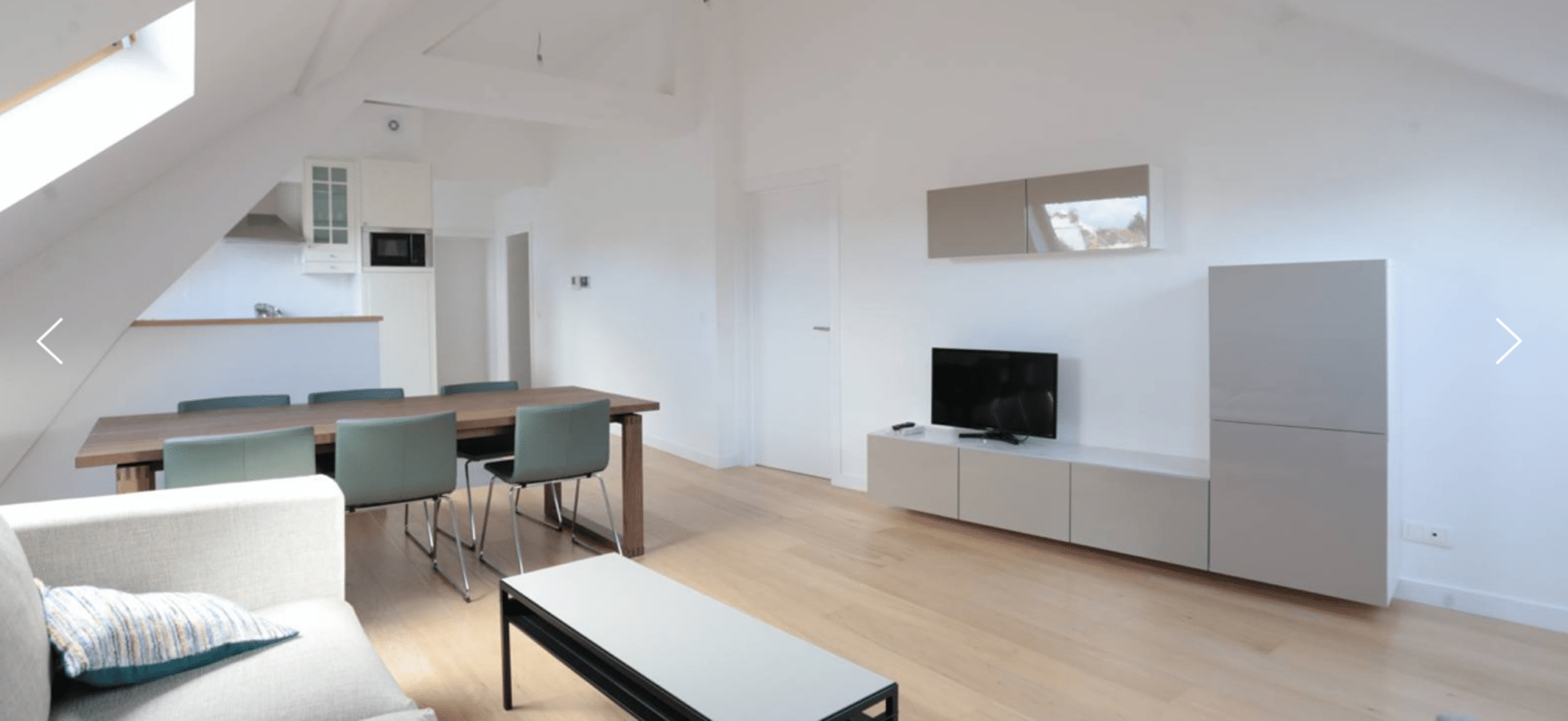 whise photo 3 Open-plan living space with a wooden dining table and teal chairs, white walls, and a TV cabinet on the right.