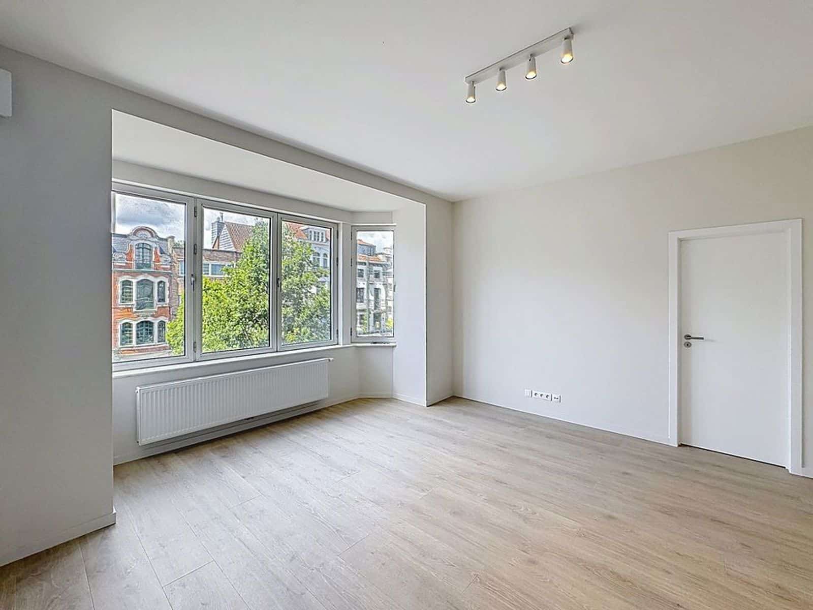 Salon Lumineux avec Parquet Clair – You Real Estate Agency Salon lumineux rénové avec parquet clair, grandes fenêtres et radiateur.
