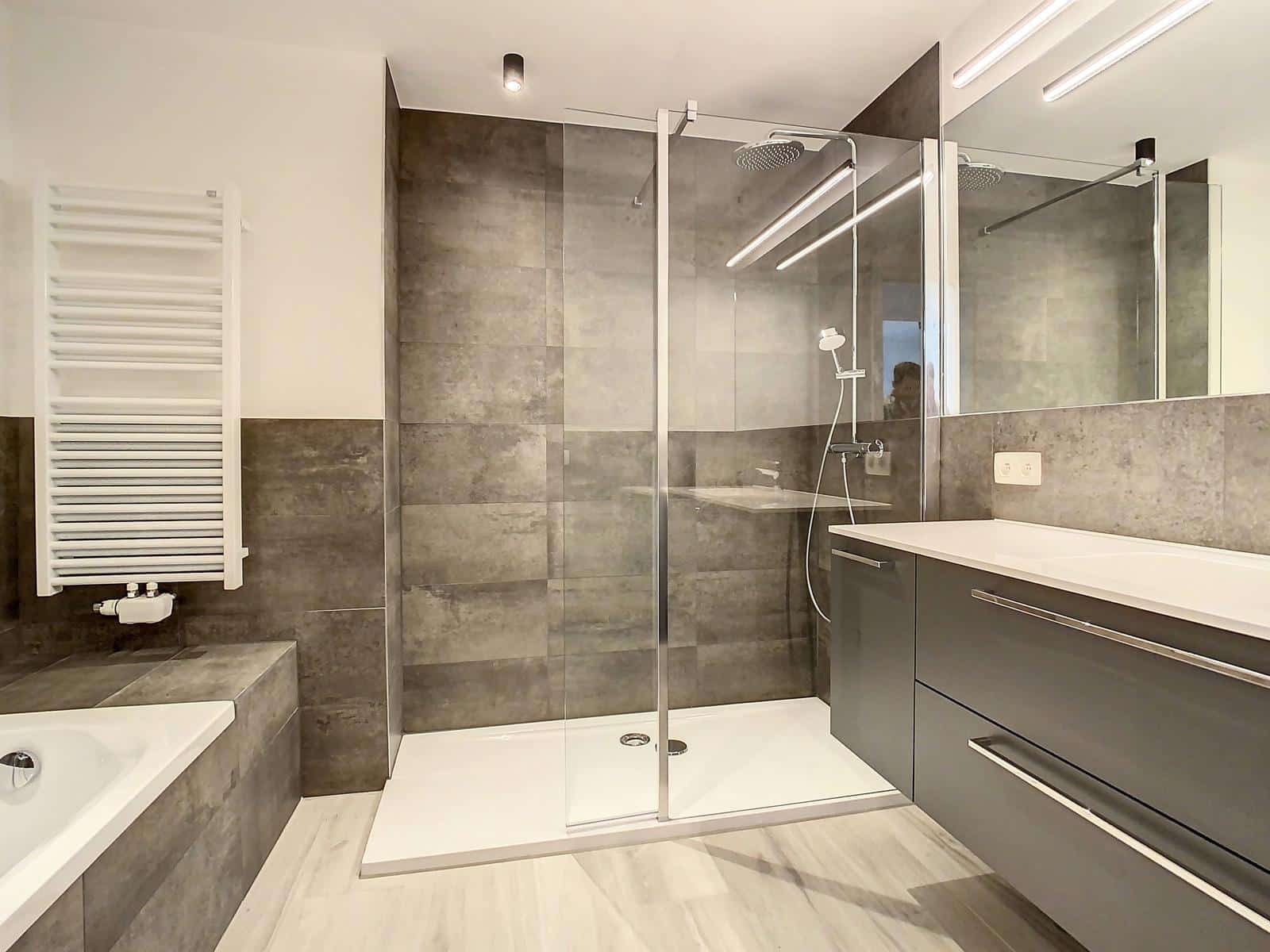 whise photo 10 A modern bathroom with a glass shower enclosure, dark gray tiles, a white bathtub, and a gray vanity.