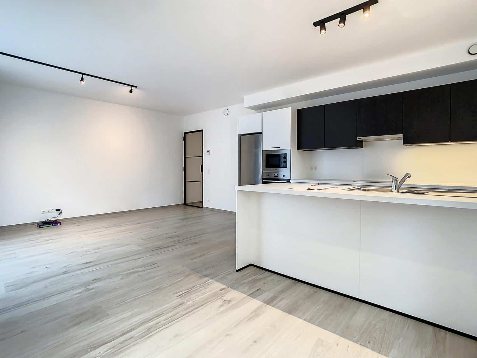 whise photo 2 Open-plan kitchen and living area with white walls, light wood flooring, black upper cabinets, a stainless steel refrigerator, a built-in oven, and a white island with a sink (empty).