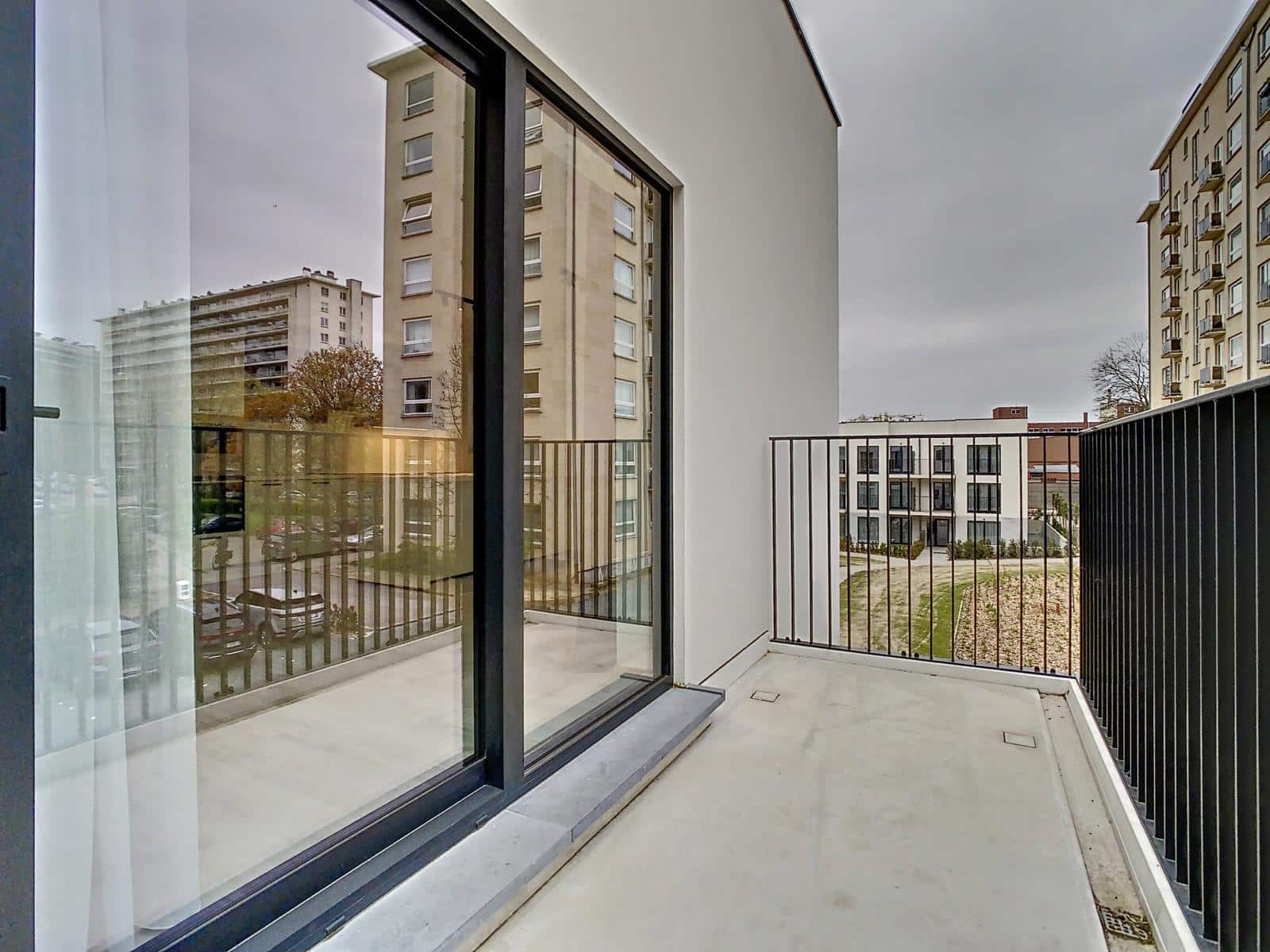 whise photo 4 A modern apartment balcony with a sliding glass door, metal railing, and a view of neighboring high-rise buildings and a courtyard outside.