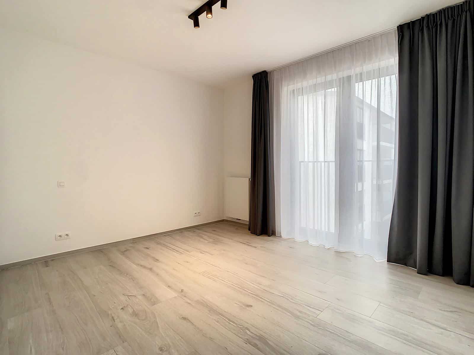 whise photo 6 An empty, modern room with light wood flooring, white walls, and a large window covered by sheer white and dark curtains