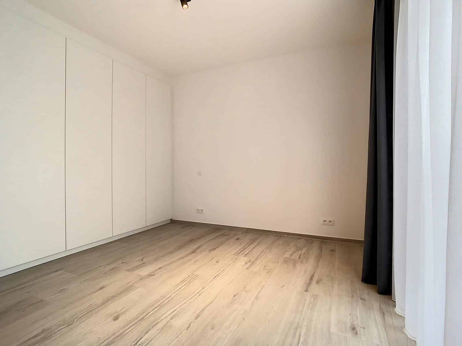 whise photo 7 An empty room with white built-in closets along the left wall, light wood flooring, and a large window with white and dark curtains on the right.
