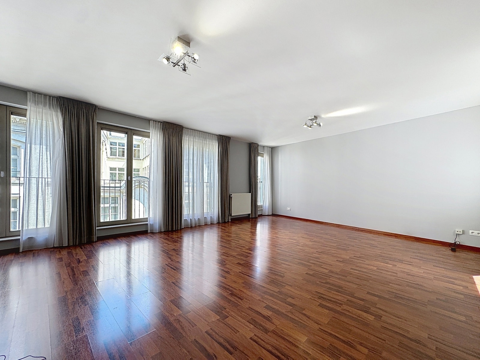 whise photo 2 Empty modern living room with dark wood floors, large windows with sheer and dark curtains, and ceiling spotlights.
