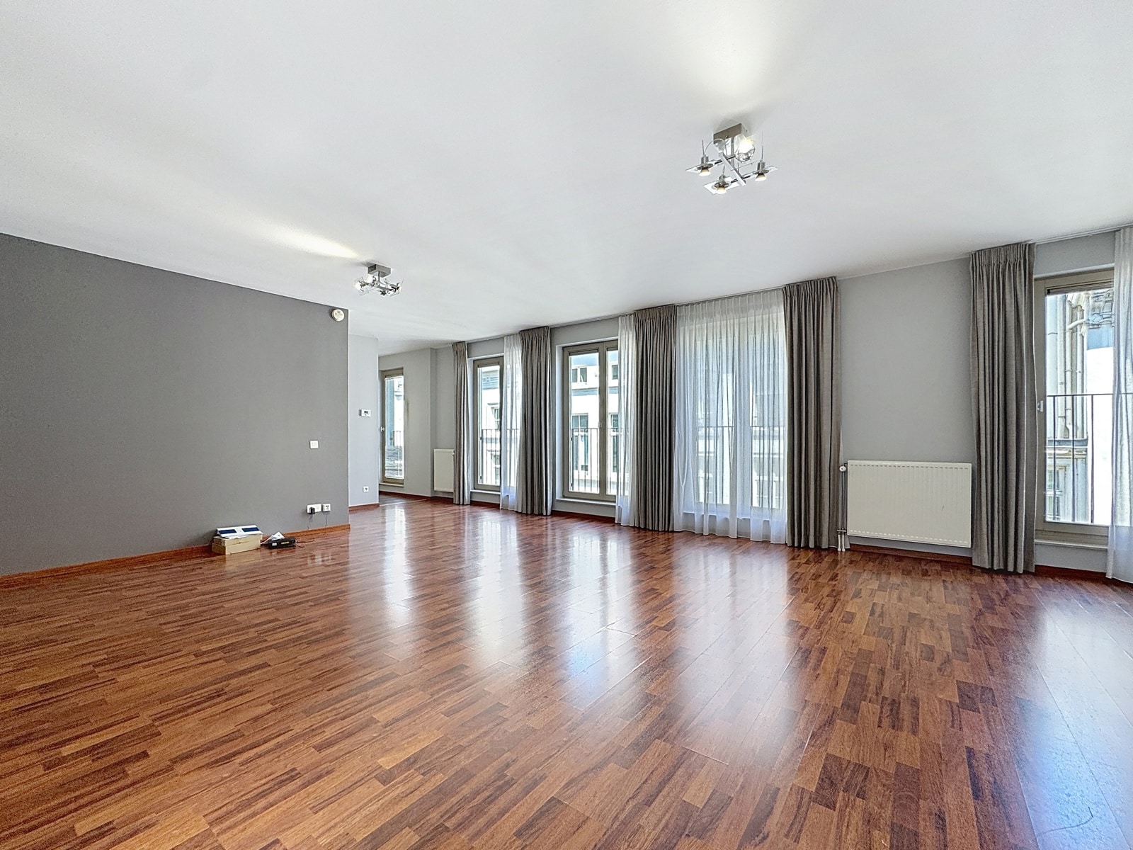 Salon Lumineux avec Baies Vitrées – You Real Estate Agency Grand salon d’appartement lumineux avec parquet, baies vitrées et rideaux gris.