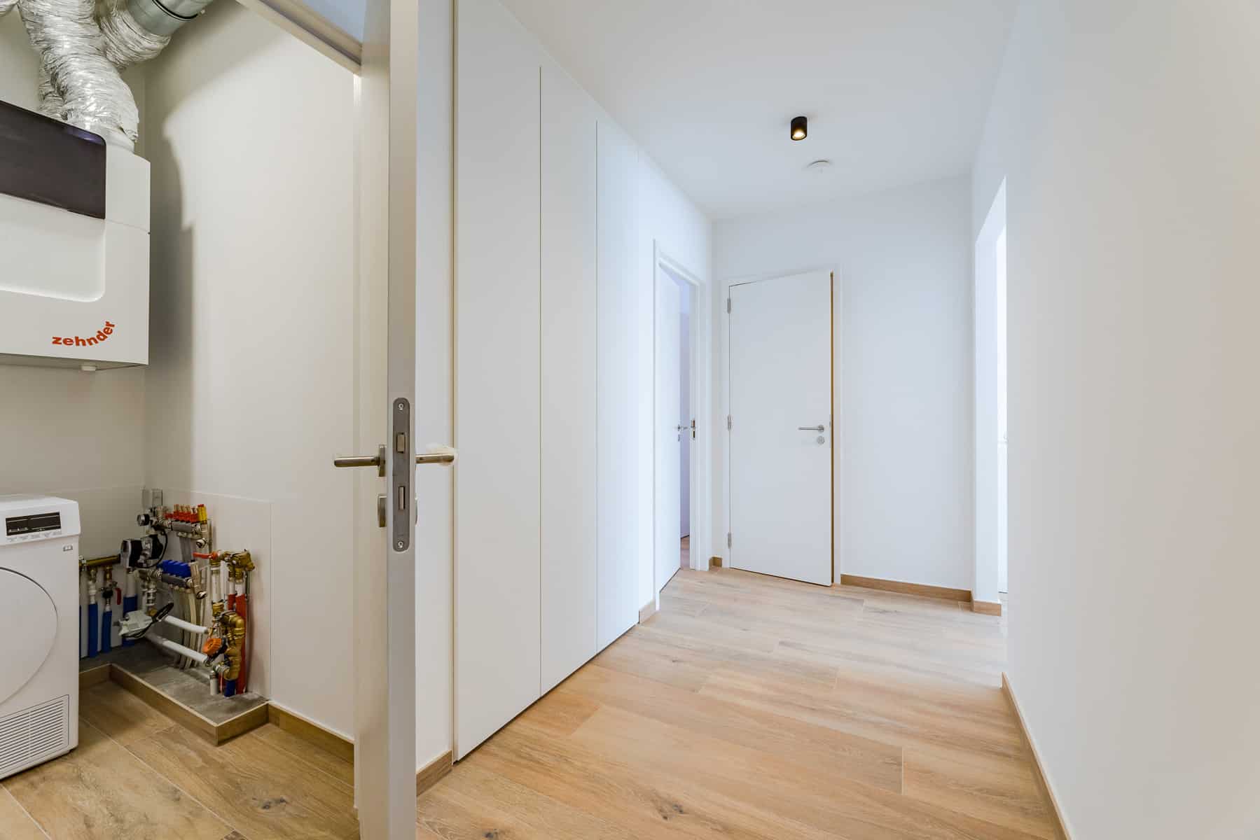 Moderne gang met wasruimte – You Real Estate Agency Moderne gang in een appartement met inbouwkasten en een technische wasruimte.