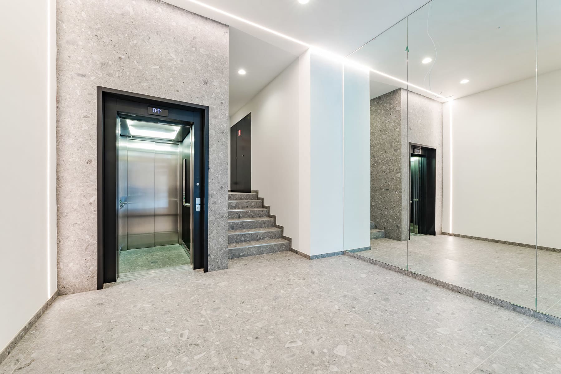 Wijze foto 11 Moderne lobby met een open metalen liftdeur in grijs terrazzo, naast een grijze stenen trap en een spiegelwand.