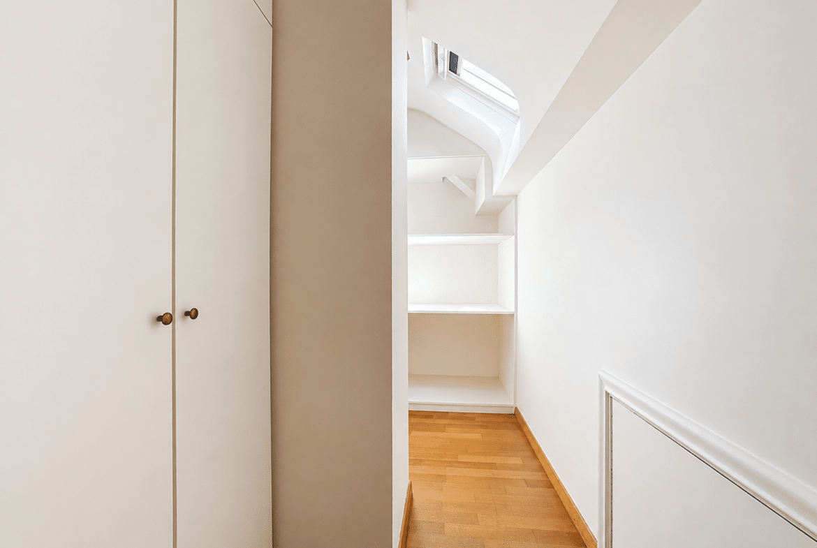 Minimalist white hallway with built-in shelves at the end and a wooden floor; staircase visible to the right.