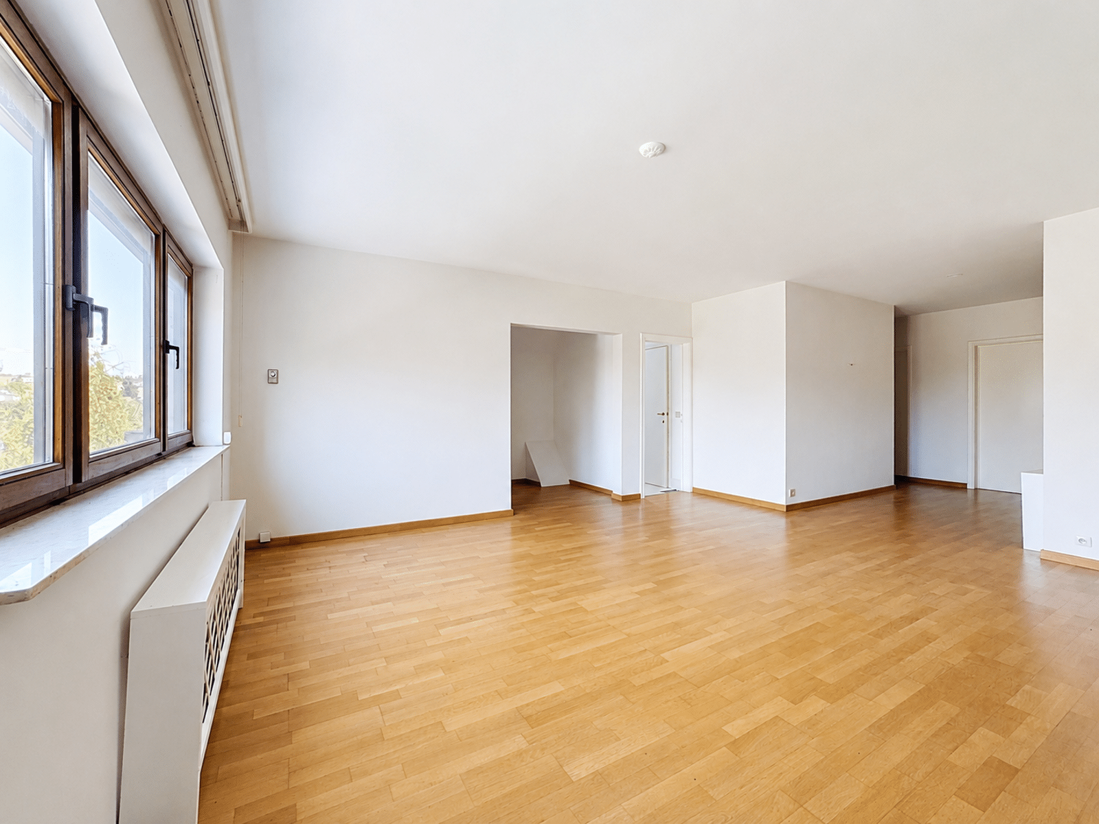Salon Lumineux avec Grandes Fenêtres – You Real Estate Agency Salon lumineux d’appartement vide avec parquet et grandes fenêtres.