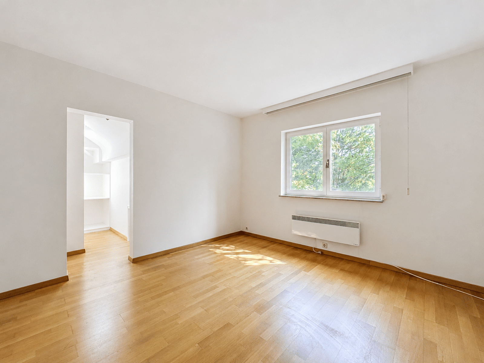 whise photo 7 Empty sunlit living room with light wood flooring, white walls, a large window, and a wall-mounted heater beneath the window; doorway to a small closet or alcove on the left.