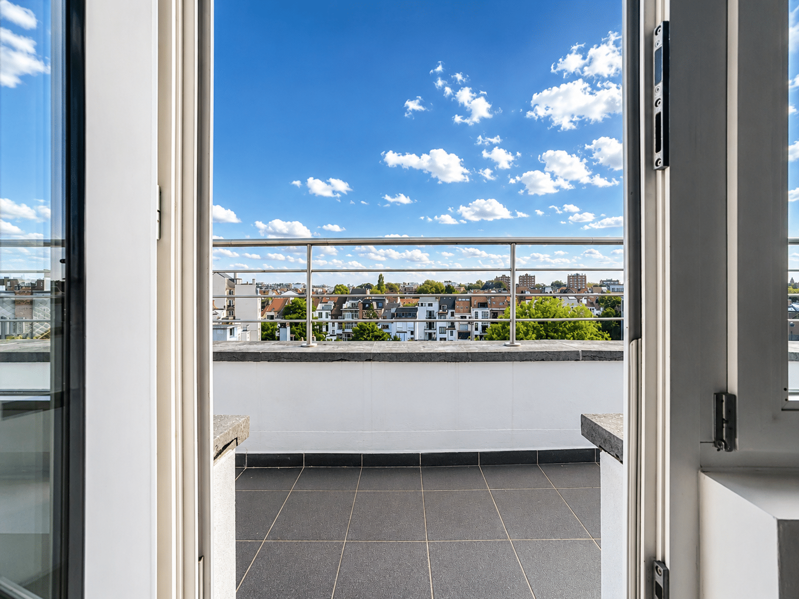 whise photo 14 Open balcony door/frame looking out over a tiled terrace with a metal railing to a cityscape and blue sky with scattered clouds.