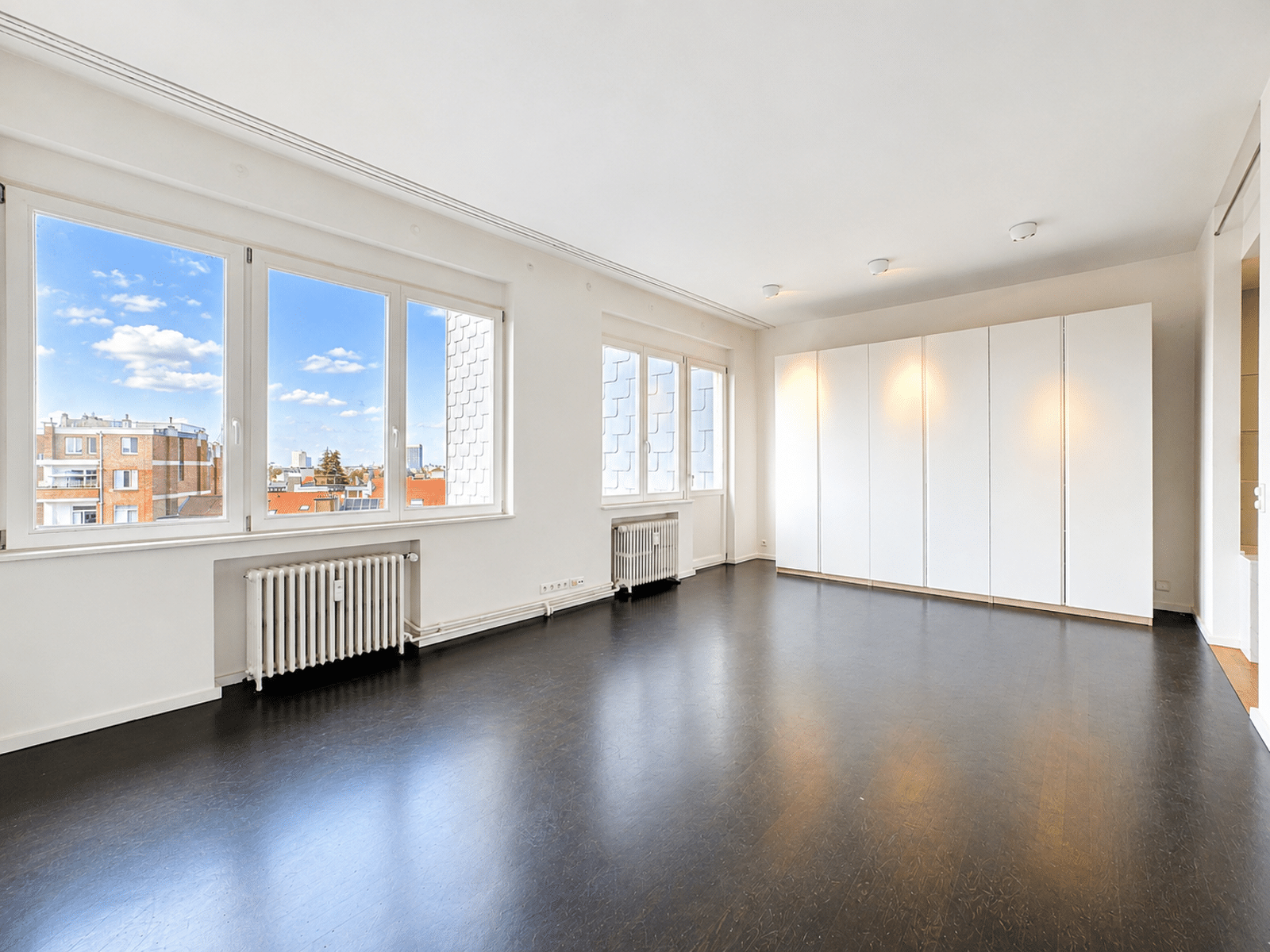 Salon Lumineux avec Placards Intégrés – You Real Estate Agency Salon lumineux d’appartement avec parquet foncé, grandes fenêtres et placards intégrés.
