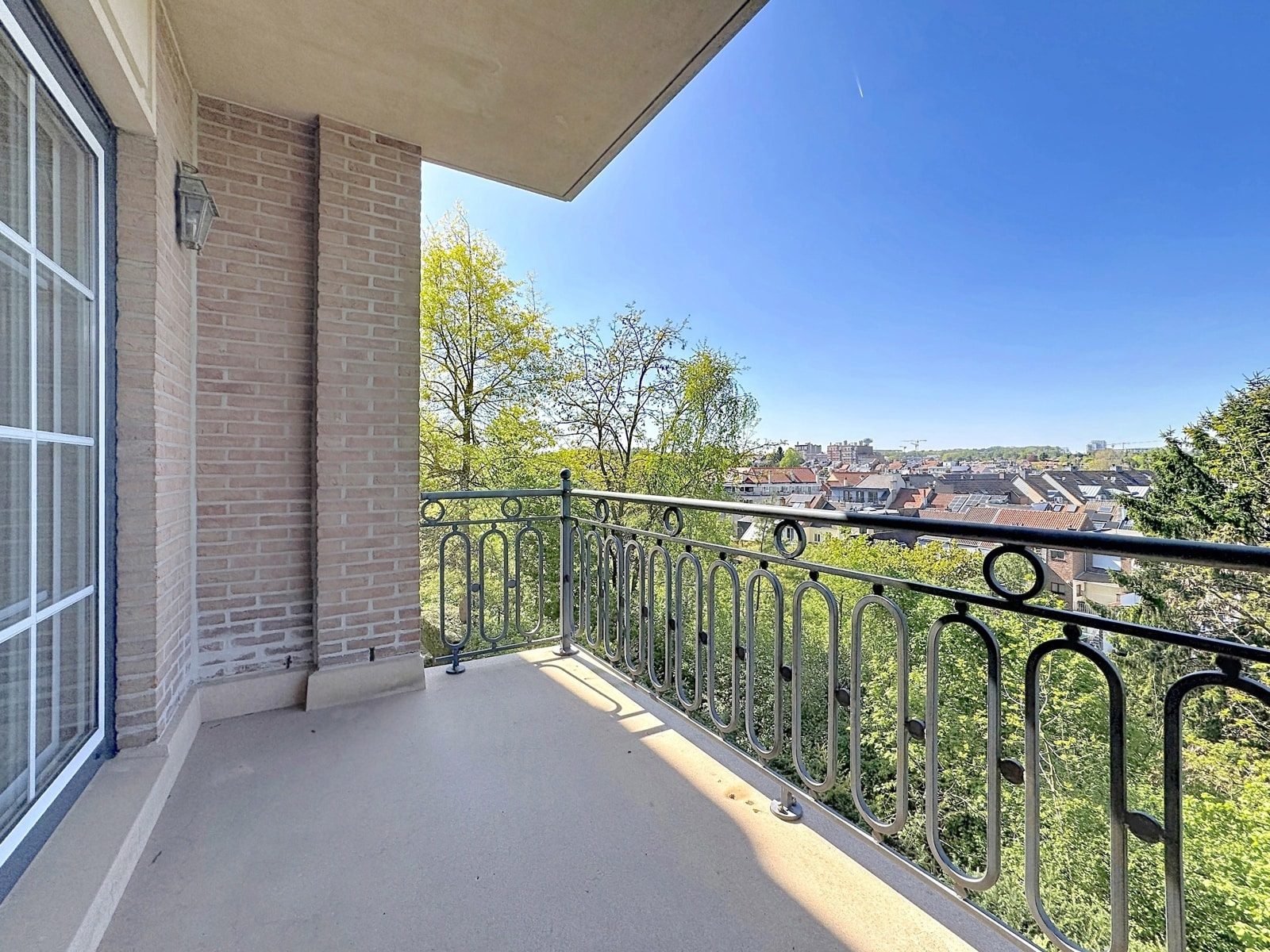 whise photo 4 Balcony with ornate metal railing overlooking green trees and a city neighborhood under a clear blue sky.