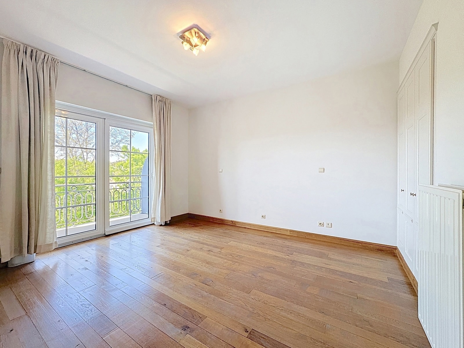 whise photo 6 Empty sunlit room with wooden floor, white walls, and a built-in white wardrobe on the right, plus glass balcony doors with curtains.
