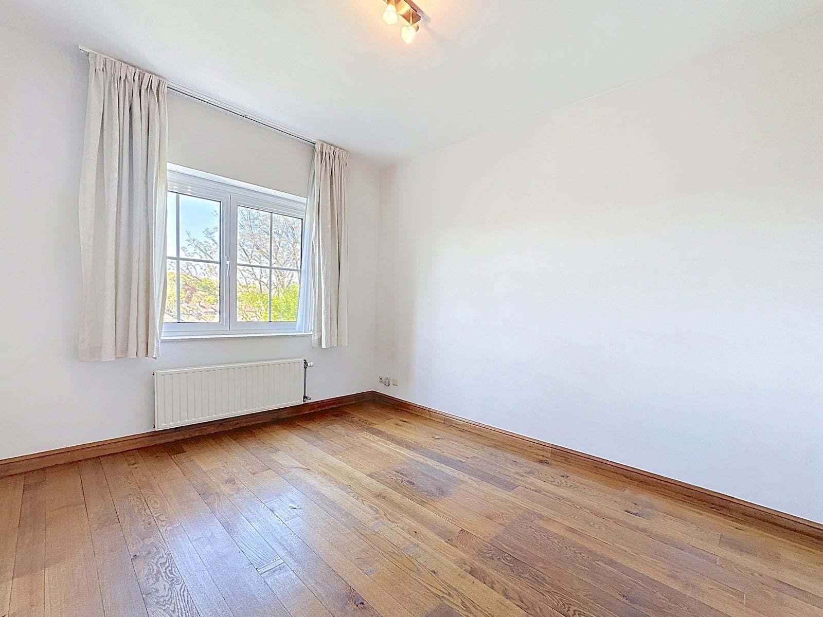 Chambre Lumineuse avec Parquet – You Real Estate Agency Chambre lumineuse d’appartement avec parquet, fenêtre, rideaux et radiateur.